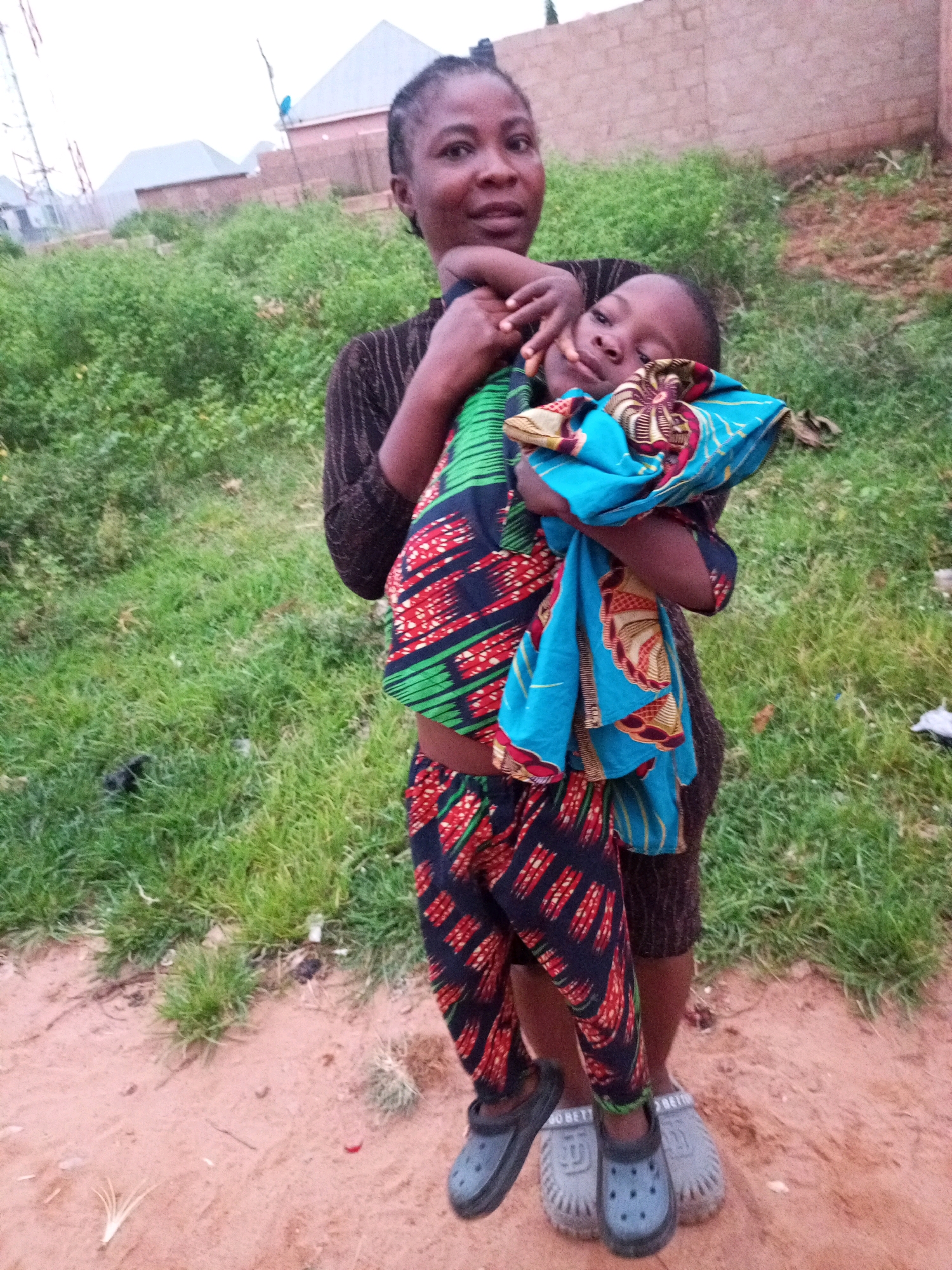 The image depicts a woman holding a young child in her arms outdoors. Both are wearing vibrantly patterned clothing. The woman is dressed in a dark, possibly brown or black, patterned top and a colorful wrap skirt or pants featuring green, red, and black designs. The child is swaddled in a bright blue fabric with red and yellow accents, and also appears to be wearing patterned pants that match the mother's. They are standing on a dirt path with sparse green grass and some dried vegetation in the background. Beyond the vegetation, there are hints of buildings, including what appears to be a brick wall and the roofs of houses, suggesting a semi-urban or rural setting in Tempure, Nigeria. The lighting suggests it is either early morning or late afternoon. The woman is looking towards the camera, while the child's face is partially obscured by the fabric, looking in a similar direction. The overall impression is a moment of familial connection captured in an everyday environment.