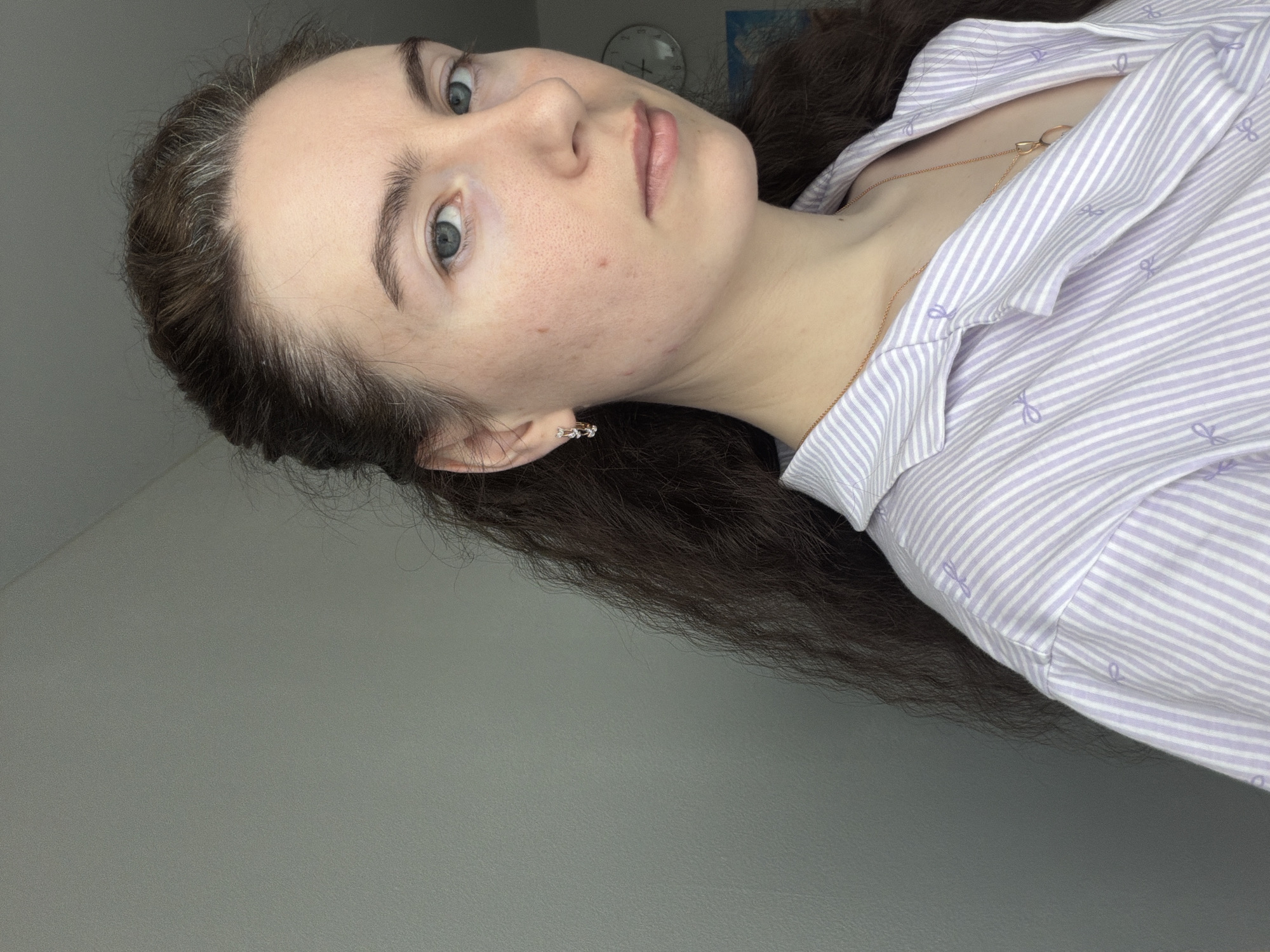A young woman with fair skin and blue eyes is depicted in a close-up portrait. She has dark hair with some graying at the roots, and is wearing a light purple and white striped shirt. A thin gold necklace is visible around her neck. The background is a plain gray wall. A clock and a map are partially visible behind her. The location is not specified to be in Yanino-1, Russia.