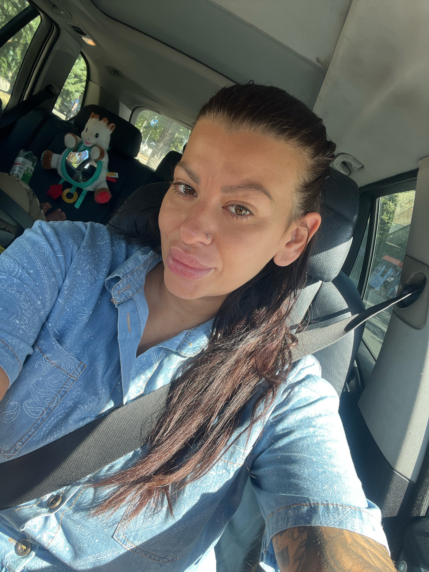In the city of Plovdiv, Bulgaria, a woman with long, dark hair pulled back in a ponytail takes a selfie from the driver's seat of a car. She is wearing a light blue denim shirt with a paisley pattern and a seatbelt. A baby toy giraffe with a mirror in its stomach is visible in the back seat behind her. Sunlight streams in through the car windows, creating shadows. The woman has a nose ring and visible tattoos on her arm. The background outside the car appears to be a street with trees and other vehicles.