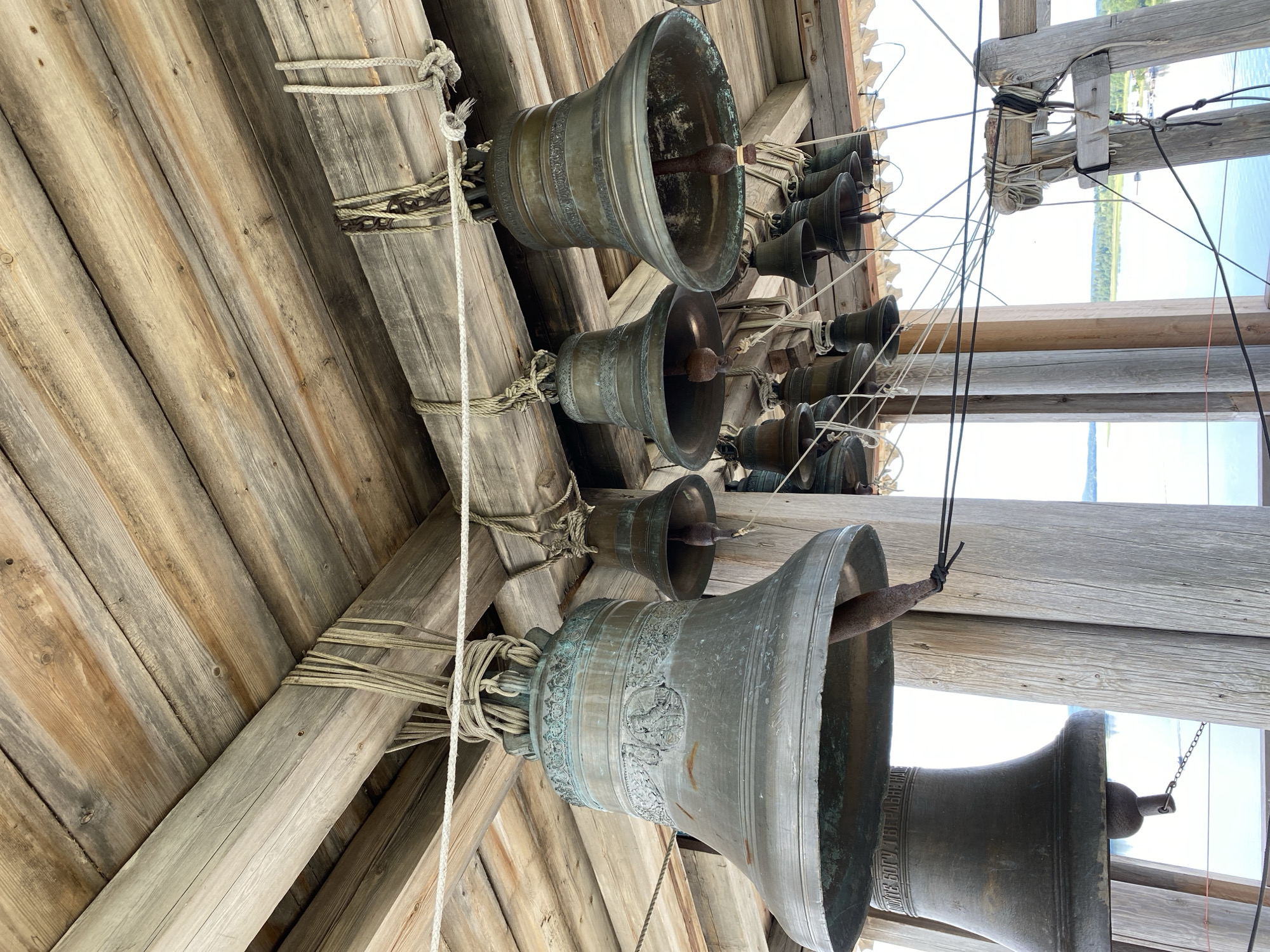 In Boyarshchina, Russia, a collection of large bronze bells are suspended from a wooden structure. The bells vary in size and are adorned with decorative bands and some with inscribed text. They hang from thick ropes tied to wooden beams, which form the roof of the structure. The wooden beams show signs of age and weathering. In the background, a body of water can be seen, with a distant shoreline dotted with trees. The overall scene suggests a bell tower or a belfry.