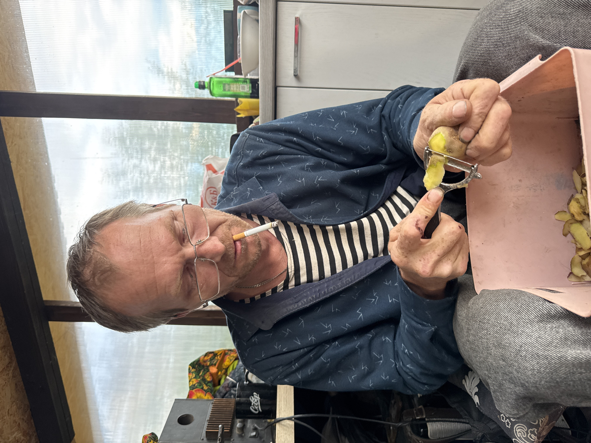 A man is peeling a potato with a vegetable peeler. He is seated and wears glasses, a striped shirt, and a blue jacket. He has a cigarette in his mouth. A pink container sits next to him, filled with potato peelings. The background shows a window with outdoor light and a glimpse of a cabinet. Below the man's arm is an electric tuner. The scene is likely indoors. The location is Chaikovsky, Russia.