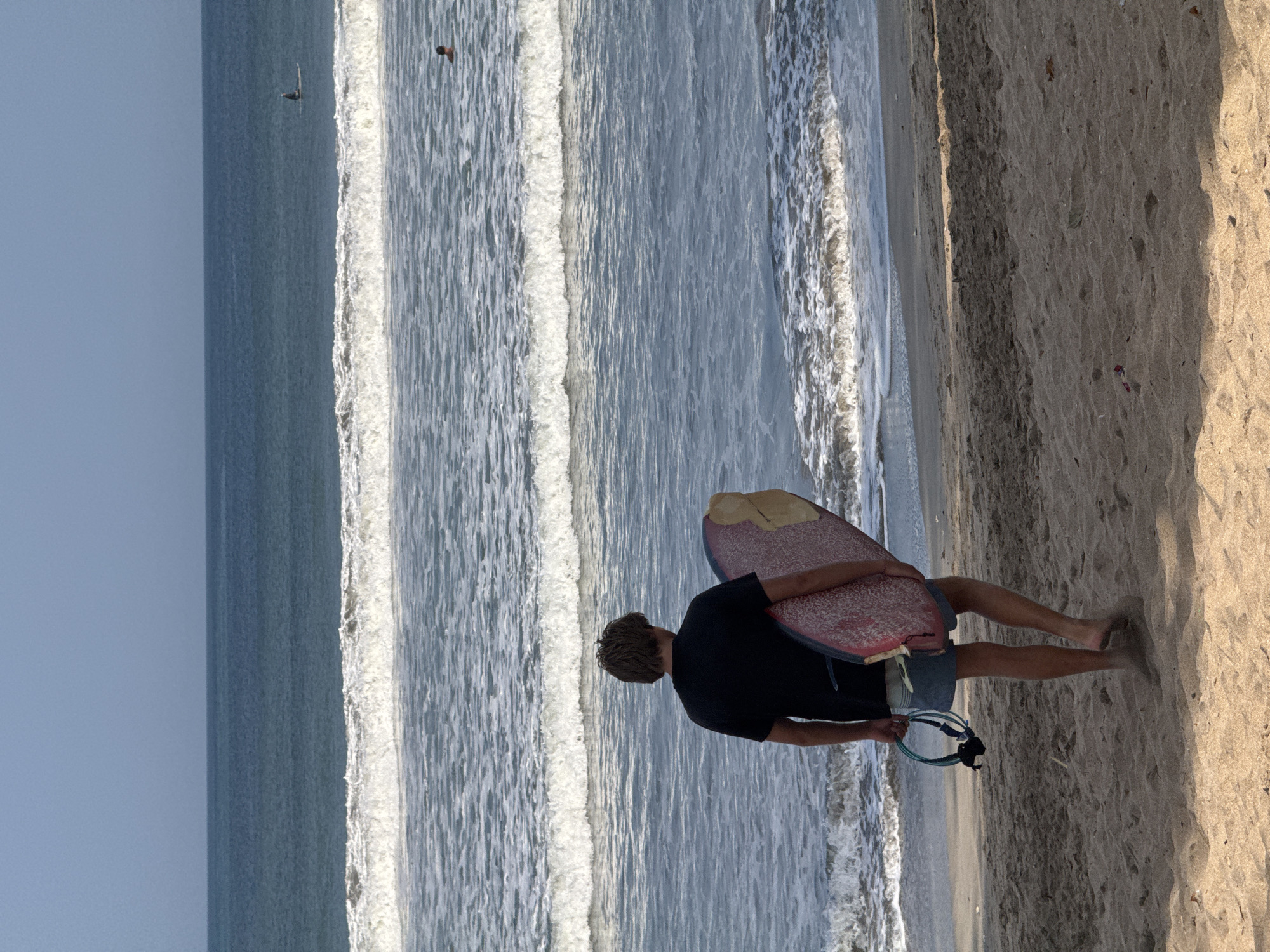 A young man with brown hair and a black t-shirt walks away from the camera on a sandy beach, holding a surfboard under his right arm. He is wearing blue shorts and carrying a surf leash in his left hand. The ocean is calm with small waves rolling onto the shore, and a few people can be seen in the water in the distance. The sky is clear and blue. The scene appears to be a typical day at the beach in Kuta, Indonesia, a popular surfing destination.