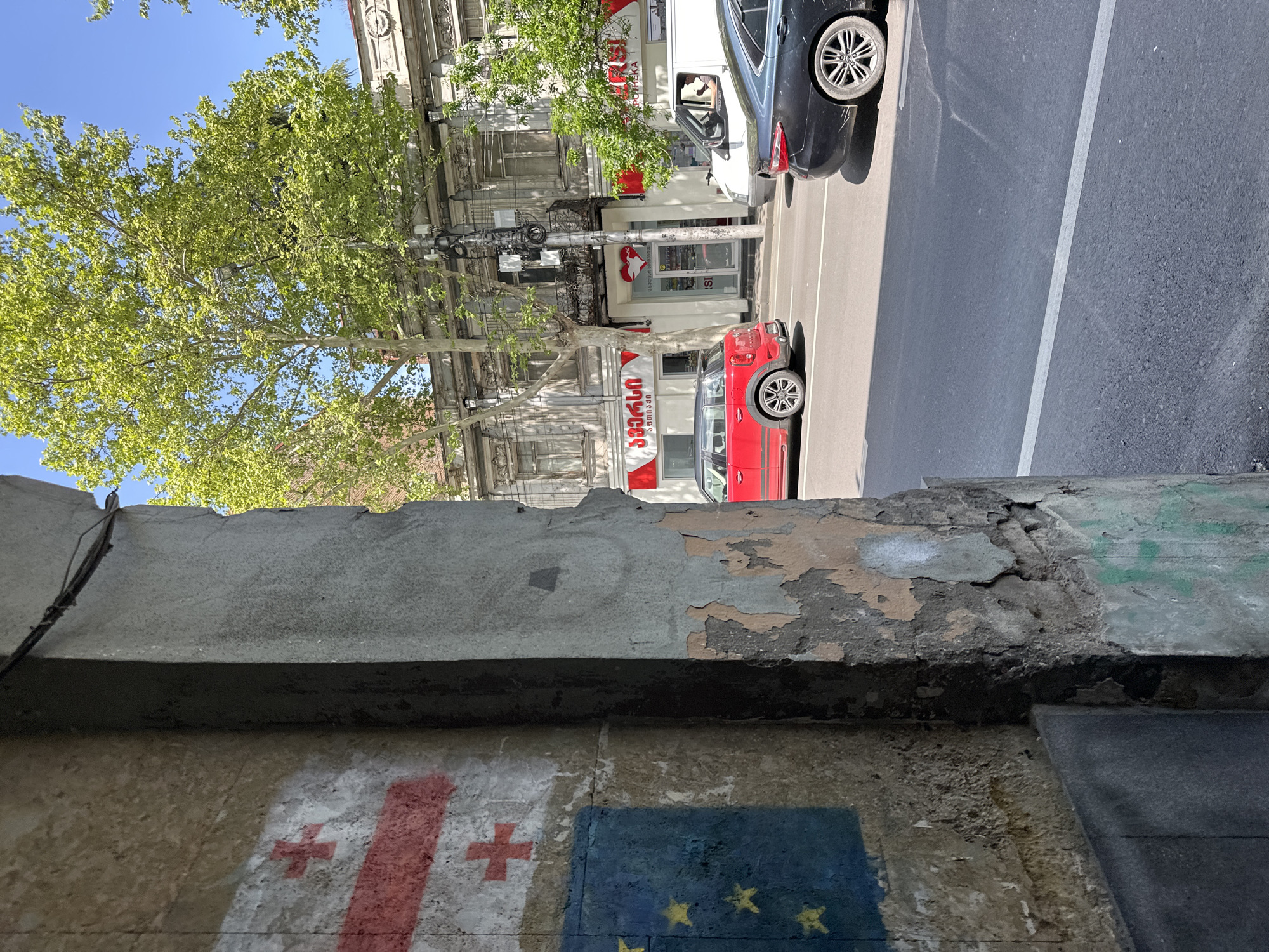 The image shows a street scene in Tbilisi, Georgia, viewed from behind a weathered concrete structure adorned with painted flags: the Georgian flag (white with red crosses) and the European Union flag (blue with yellow stars). The street is lined with old buildings with architectural details. A tree with green leaves partially obscures the view. Cars are parked along the street, including a red car and a white van. A pharmacy with a red heart logo is visible in the background. The asphalt road has a white lane marking. The sky is visible in the upper left corner.