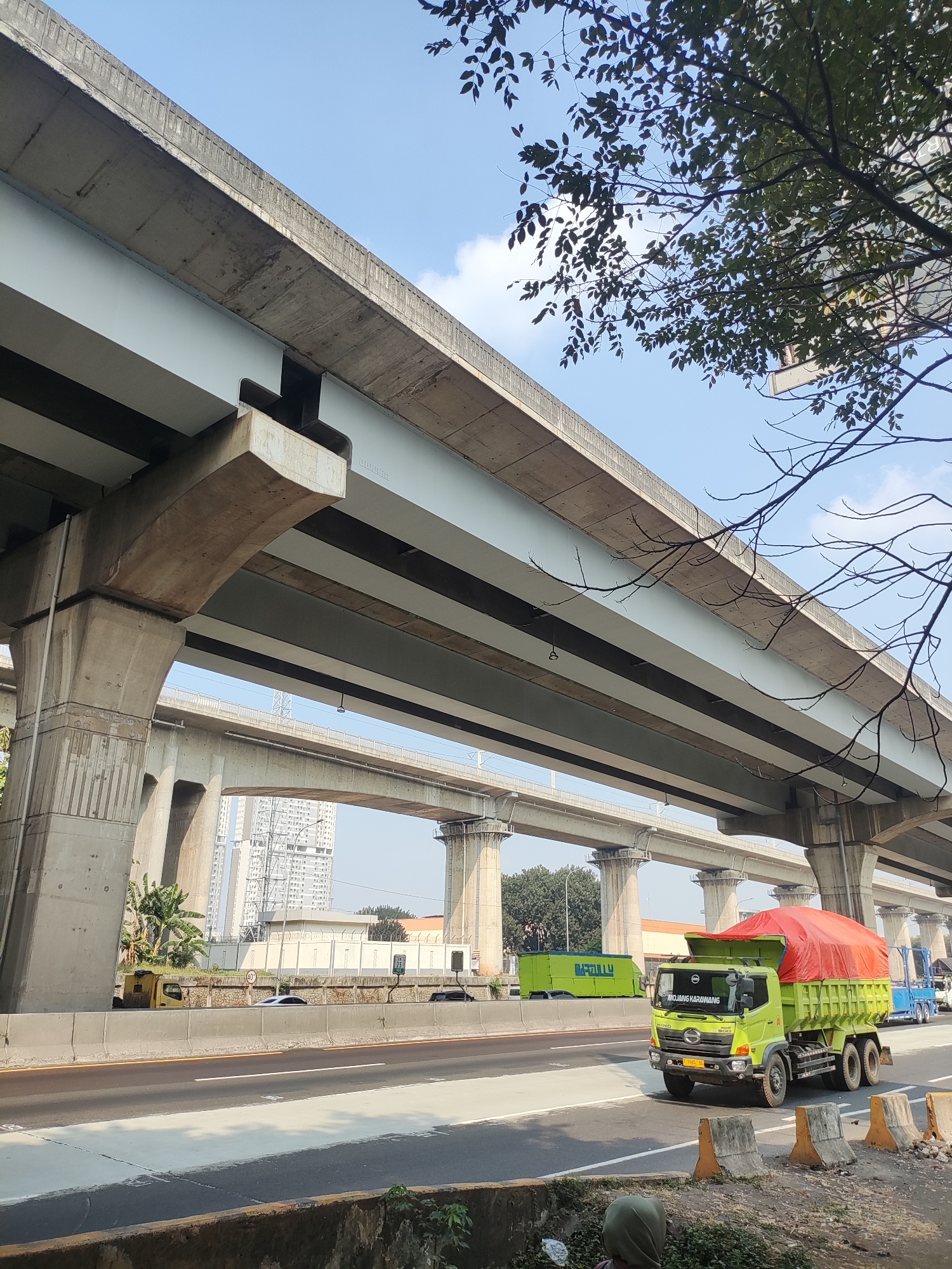 In Lambangsari, Indonesia, a bright green dump truck with a red tarp covering its load drives on a multi-lane highway beneath an elevated concrete overpass. The truck, a Hino 500 series, has visible text on its windshield that reads "MJOJANG KARAWANG". In the distance, other vehicles, including a blue truck and a white car, travel on the highway. The scene is framed by the overpass's massive concrete pillars and a lush green tree with dark leaves. The sky above is clear blue with a few white clouds. The road is asphalt with white lane markings and a light gray painted section. Concrete barriers and traffic cones line the side of the road. In the background, tall buildings suggest an urban environment.