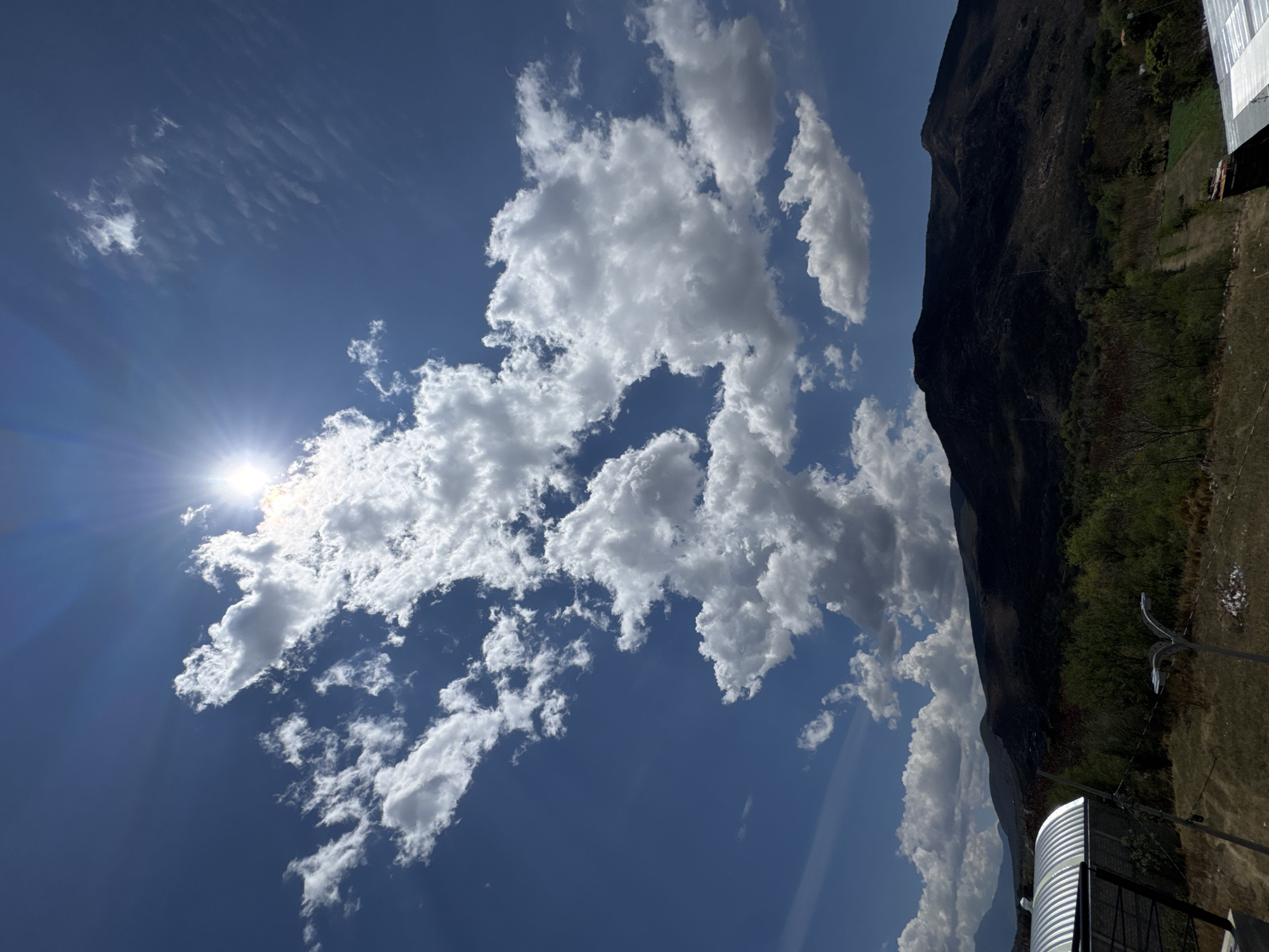 The image depicts a daytime scene in Tlalixtac de Cabrera, Mexico. A bright sun shines from the upper left, partially obscured by scattered, puffy white cumulus clouds against a vibrant blue sky. The right side of the image shows a dark, steep mountainside covered with vegetation. In the lower right corner, there's a structure with corrugated metal, along with utility poles and a grassy area. The overall impression is one of a bright, sunny day with a mix of natural and built environments.