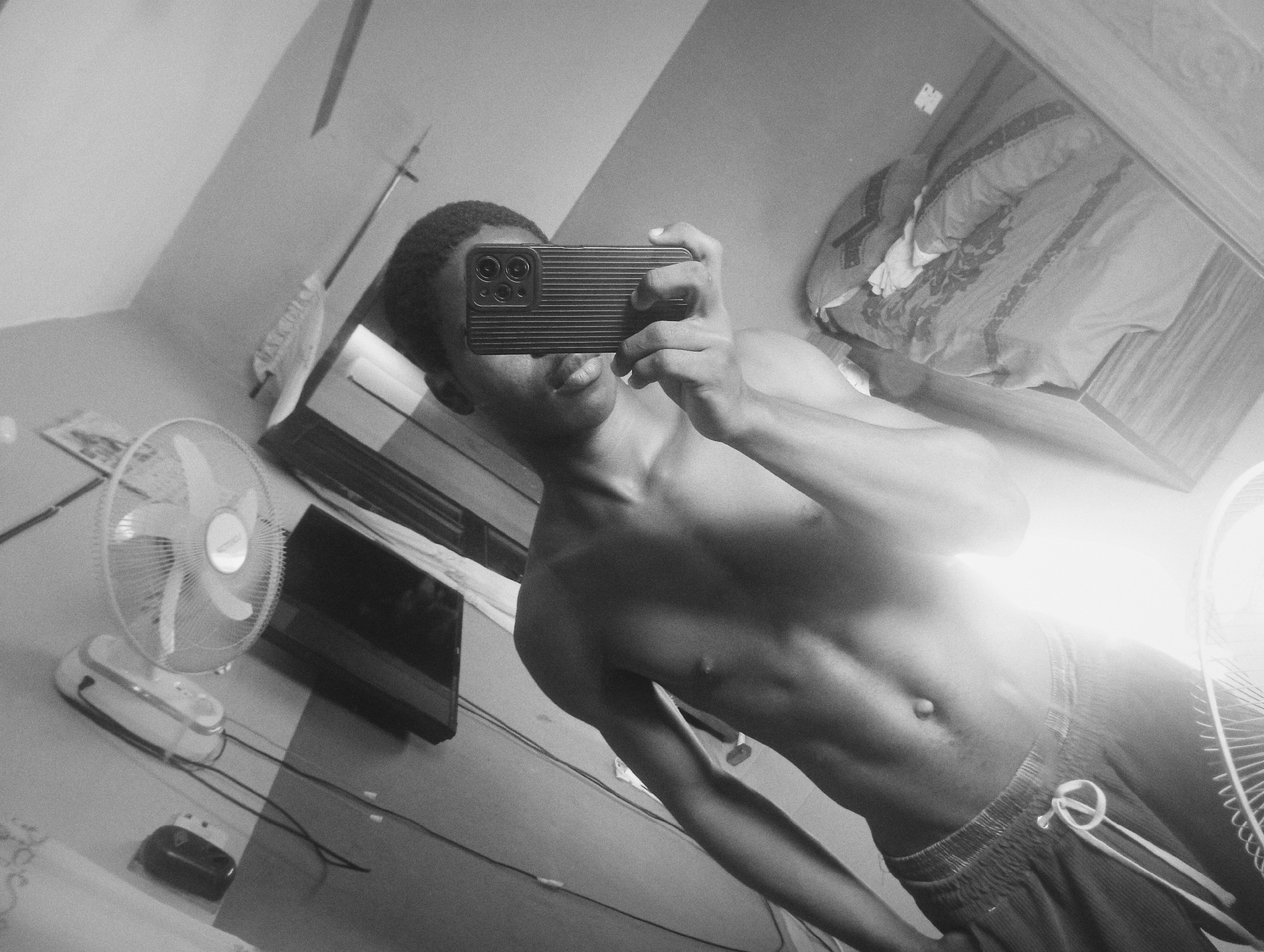 A young individual, likely male, takes a shirtless mirror selfie in what appears to be a bedroom in Nkpor, Nigeria. The image is rendered in black and white with a noticeable grainy texture. The subject, seen from the waist up, holds a dark-cased smartphone with multiple camera lenses to obscure their face, revealing only their chin and mouth. They have short, dark hair and a lean physique with visible abdominal muscles.
The setting is an indoor room, characterized by light-colored walls. Reflected in the mirror on the upper right, a bed with rumpled patterned bedding is visible. In the lower-left, a white oscillating stand fan rests on a surface, next to what appears to be another white electronic device with black wires, possibly a power strip or adapter. A dark, flat-screen television or monitor is partially visible behind these items. The person is wearing dark shorts or sweatpants with a white drawstring tied in a loop.
The photograph is taken at an upward, diagonal angle, giving a dynamic perspective. There is a bright light source in the lower-right quadrant, creating a lens flare or strong highlight on the subject's side and the fan, suggesting either strong natural light or an artificial light source. No discernible text is visible. The presence of a fan is consistent with the warm climate often found in Nigeria.
