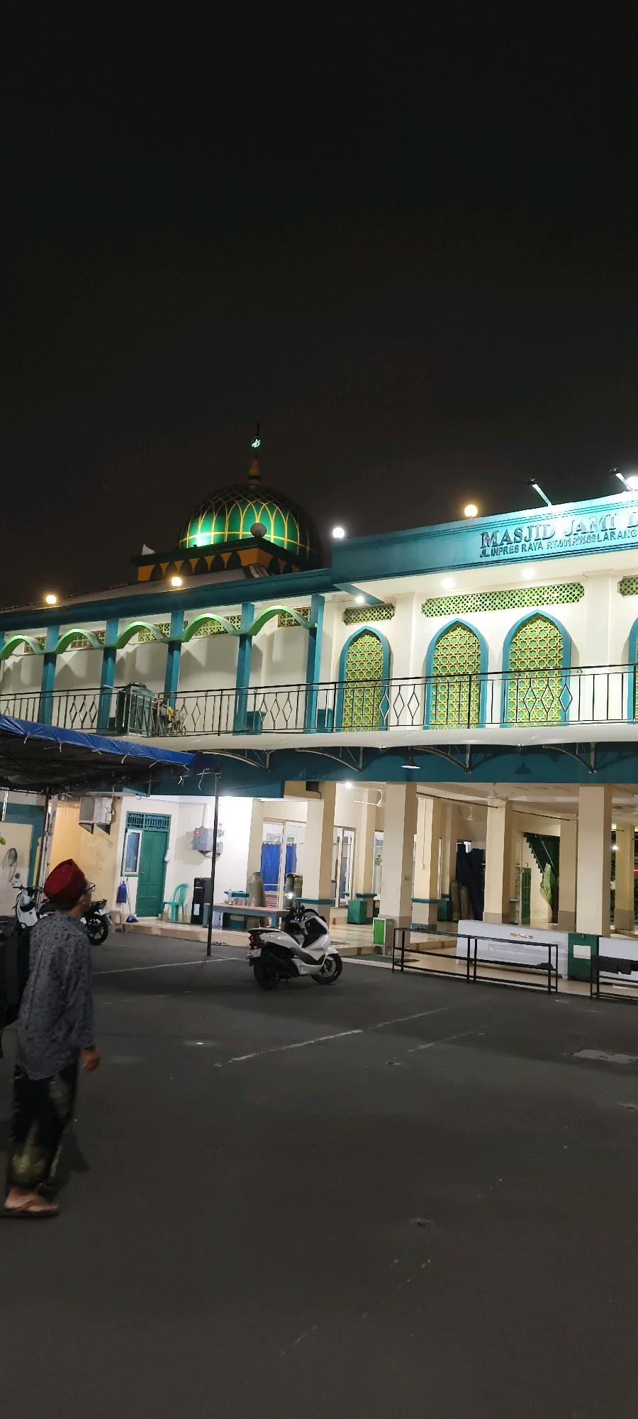 The image depicts the exterior of a mosque at night, likely in Tangerang, Indonesia, as indicated by the text on the building. The mosque features a prominent green dome with yellow accents and decorative latticework on its windows. A balcony with blue and white railings runs along the upper level. In the foreground, a man wearing a red cap and a patterned shirt walks away from the camera, his back to the viewer. Several motorcycles are parked in the paved area in front of the mosque. The environment is dark, illuminated by artificial lights from the building and possibly streetlights, creating a nighttime scene. The overall impression is one of a public building at rest after hours.