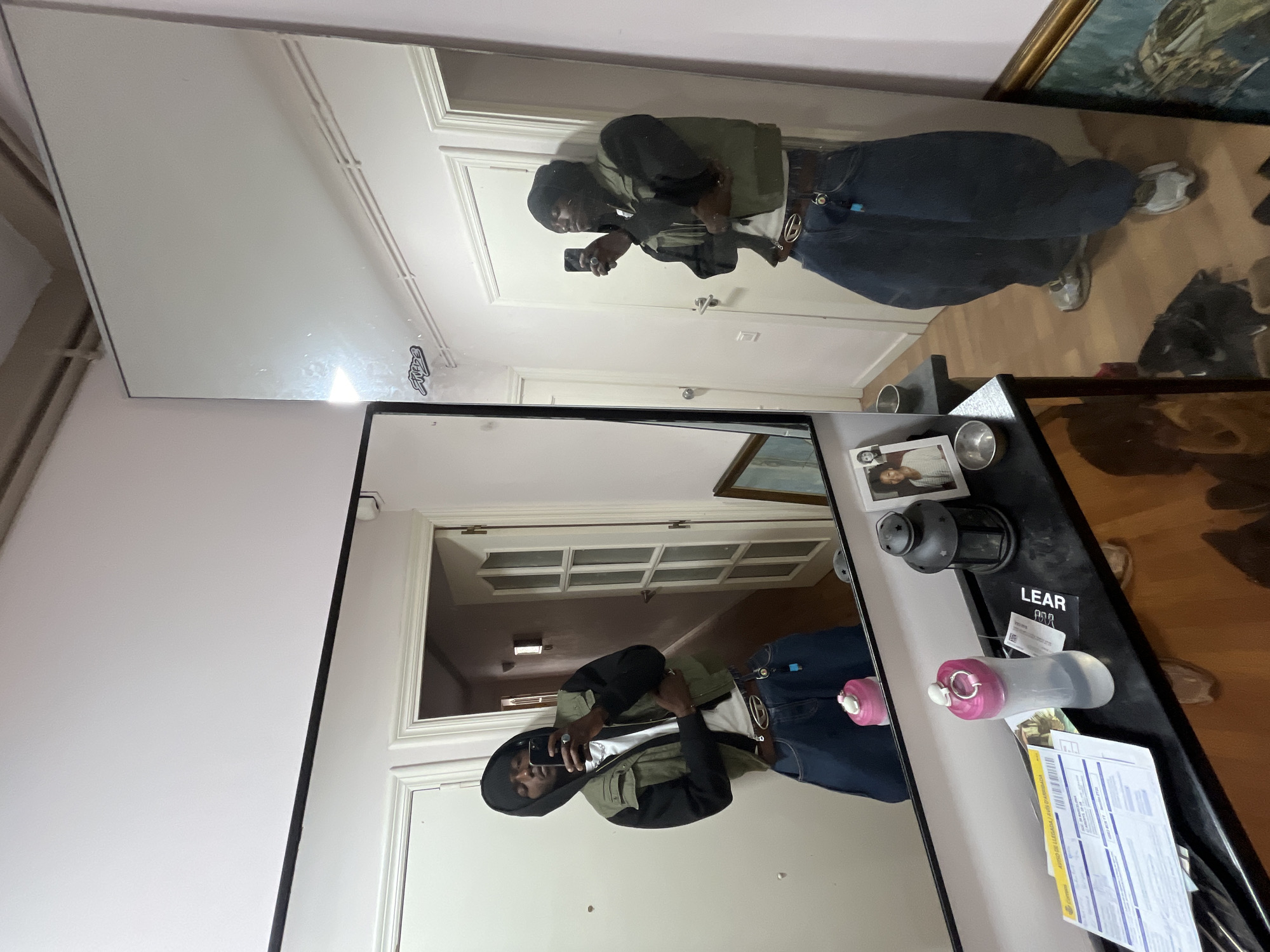 A Black male wearing a black hoodie under a khaki-colored vest, blue jeans with a large belt buckle, and grey sneakers takes a mirror selfie with his phone. The reflection is visible in two adjacent mirrors mounted on a light pink wall. The mirrors reflect a doorway and hallway behind the subject. The mirrors sit on a black console table next to various objects including water bottles, a picture frame, a lamp, and paperwork. A framed painting hangs on the wall to the right. The location is presumed to be in Barcelona, Spain. The evidence for its location cannot be seen in the given image.