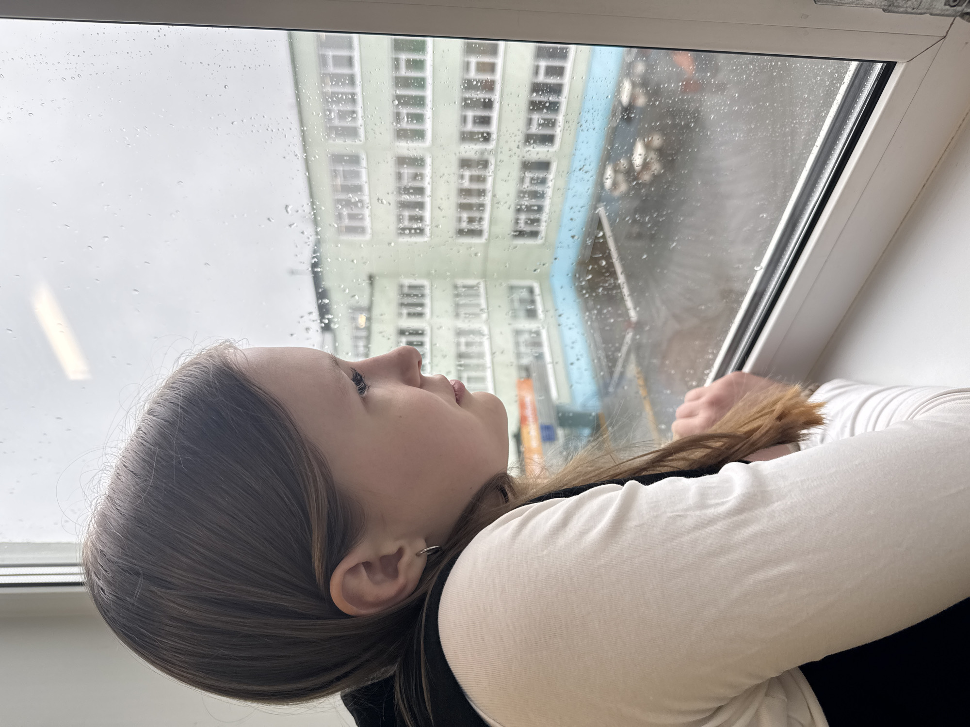 An upward-angled close-up shot captures a young person looking out a rain-streaked window. The subject, a female with long brown hair, is positioned in the foreground, her profile visible as she gazes upwards and outwards. She wears a white long-sleeved top under a darker garment. A small hoop earring is visible in her right ear.
The window pane is covered in numerous raindrops, indicating current or recent rainfall. Through the blurred, wet glass, a multi-story building with a light green facade and many rectangular windows is visible. A hint of a bright blue structure or wall is also seen on the right side of the exterior view. The sky beyond the building appears overcast and bright grey, suggesting a cloudy day. The window itself has a white frame and sill.
The person's expression appears thoughtful or contemplative as she observes the rainy outdoor scene, potentially reflecting on the weather or simply watching the world outside from an indoor vantage point. There is no visible text in the image. The scene depicts a quiet moment of observation during rainy weather in Chaikovsky, Russia.
