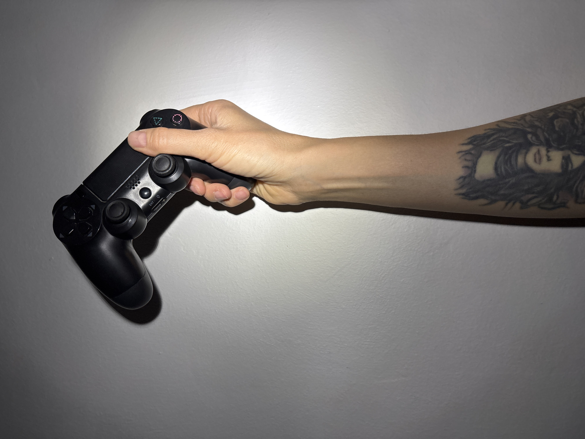 A hand with a tattoo on the forearm holds a black PlayStation 4 controller against a plain grey wall. The tattoo depicts a person's face within a complex design. The setting appears to be indoors. There is no information to suggest the location is Chaikovsky, Russia.