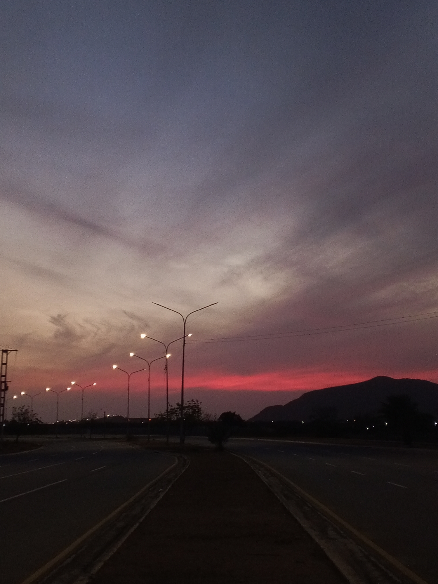 The image captures a road at dusk in Unknown, Unknown. Streetlights line the roadside, illuminating the scene with a warm glow against the backdrop of a sky transitioning from shades of purple and grey to a vibrant red-orange near the horizon. In the distance, a silhouette of a mountain range is visible. The road itself is dark, with painted lines marking the lanes. There is no visible human activity.