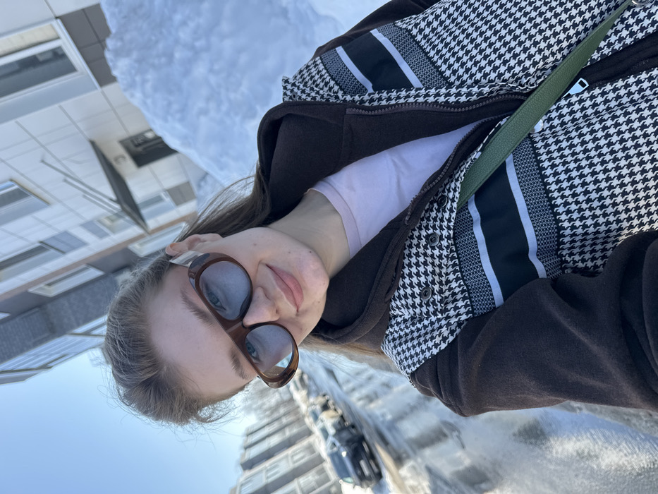 A young woman is depicted from the chest up in a bright outdoor selfie, smiling faintly directly at the camera. She wears brown oversized sunglasses with light blue tinted lenses. Her attire consists of a light pink t-shirt, a dark brown hooded jacket, and an outer garment featuring a black and white houndstooth pattern with horizontal black and light grey stripes across the chest. A dark green strap, likely from a bag, crosses her chest. Her light brown hair is visible, pulled back. The background features a clear blue sky in the upper left. Multi-story residential buildings with light-colored facades and dark windows are visible on both sides of the frame. A street with parked vehicles, including a dark SUV, runs in the mid-ground, partially covered by melting snow and slush. Large snow piles are prominent on the right side, adjacent to a building entrance with a dark overhang. The scene is brightly illuminated by direct sunlight. The location is Новое Шигалеево, Russia.
