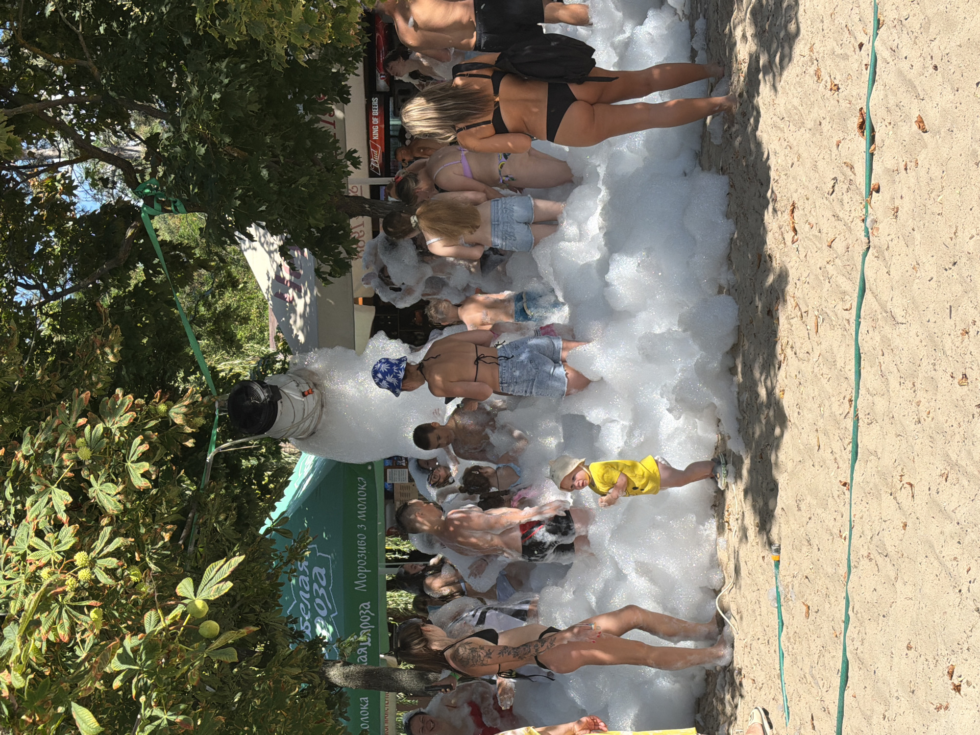 The image depicts a foam party taking place on a sandy beach, possibly in the city of Inhulets, Ukraine, based on the context provided. The scene is filled with a large amount of white foam, which obscures most of the ground and many of the people present.
Several individuals, including adults and children, are immersed in the foam. Some are wearing swimwear, while others are partially covered by the foam. A young child in a yellow shirt is standing in the foreground, looking towards the foam. In the background, there are trees providing shade, a green tent with some writing on it, and what appears to be a building with a sign that says "KING OF BEERS". A green hose is visible on the sand in the foreground, suggesting the foam is being generated by a machine. The overall atmosphere appears to be festive and casual.