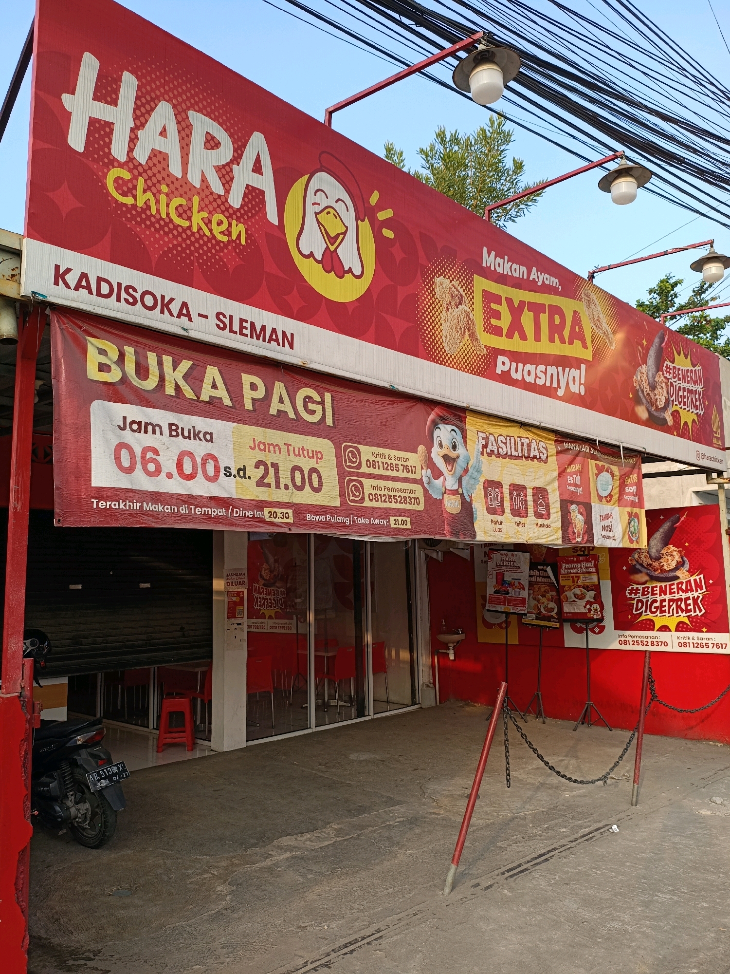The image shows the exterior of a "Hara Chicken" restaurant in Maguwoharjo, Indonesia. A large red banner with white lettering announces "HARA Chicken" and a cartoon chicken. Below this, a smaller banner displays "BUKA PAGI" (Open Morning) with operating hours from 06:00 to 21:00. The entrance to the restaurant is visible, with red chairs and tables inside, and a dark metal security shutter. A black motorcycle is parked to the left of the entrance. The facade of the restaurant is red. In the foreground, a concrete pathway is visible, with red posts and chains. The sky is blue and some trees are visible in the background.