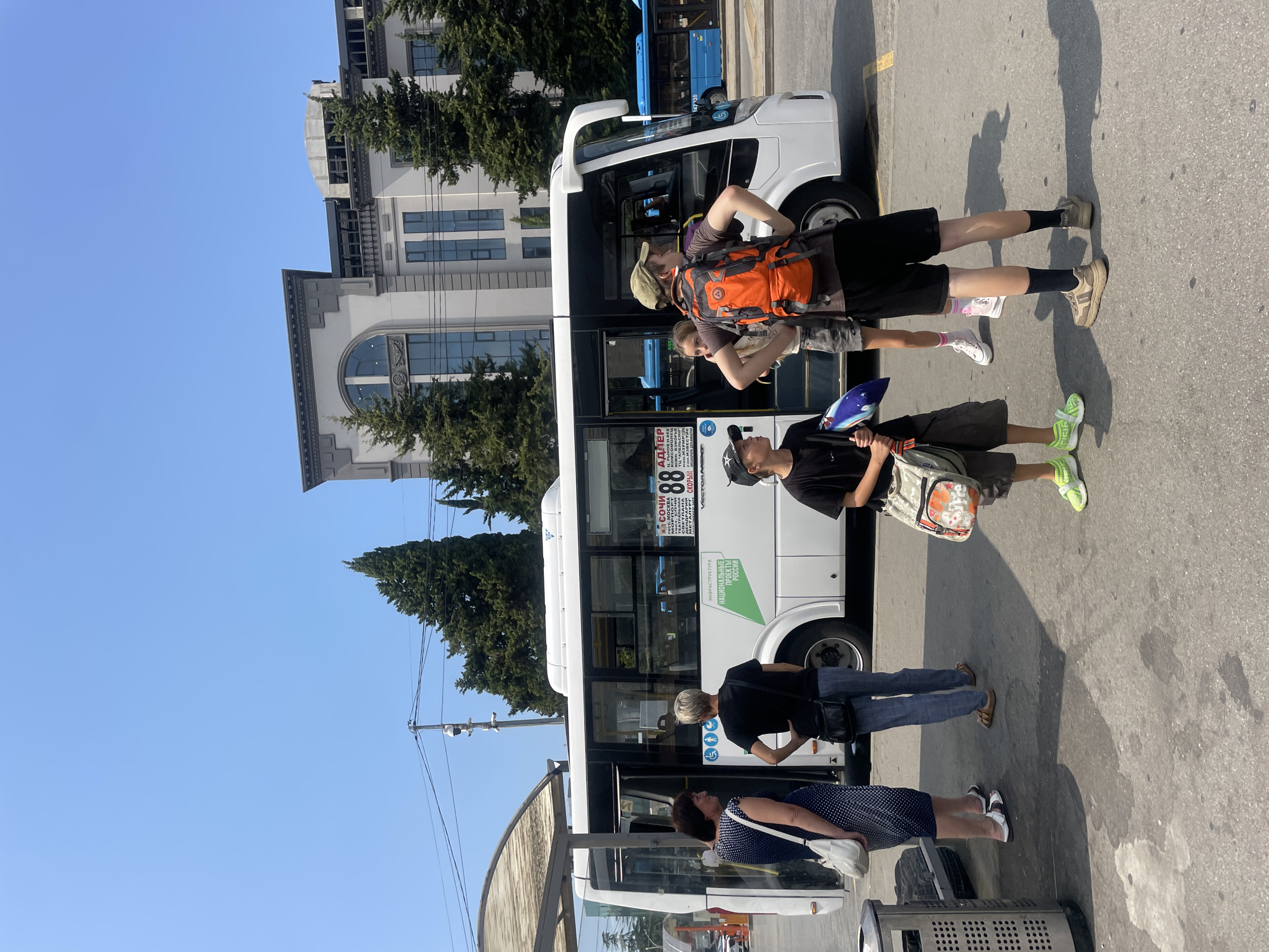 At a bus station in Sochi, Russia, a white bus with the number 88 displayed on its side is parked. A group of people stand near the bus, some boarding and others waiting. The bus stop shelter is visible on the left, and a modern building with large windows stands in the background. The sky is clear and blue, indicating a sunny day.