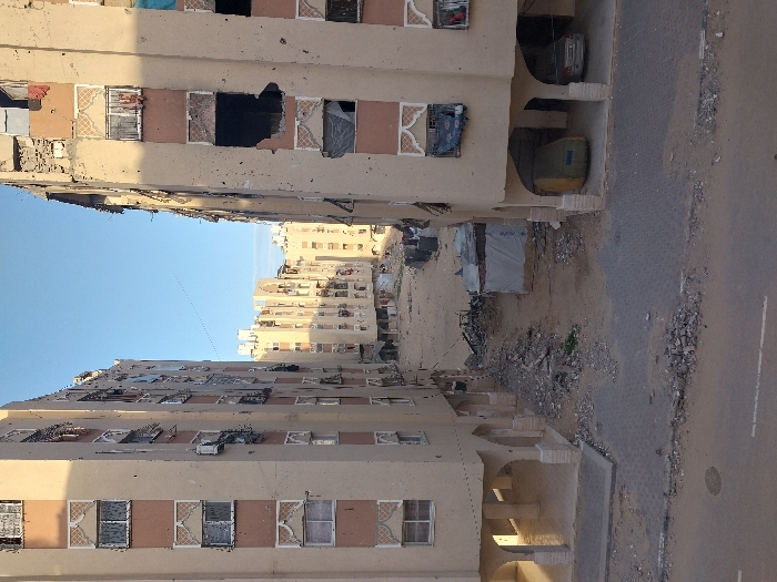 Buildings with damaged facades line a street. Windows are missing or broken, and some walls show signs of significant damage, possibly from conflict or natural disaster. Debris litters the area between the buildings. A road runs alongside one of the buildings. The buildings appear to be multi-story apartment complexes, likely located in an urban or densely populated area.