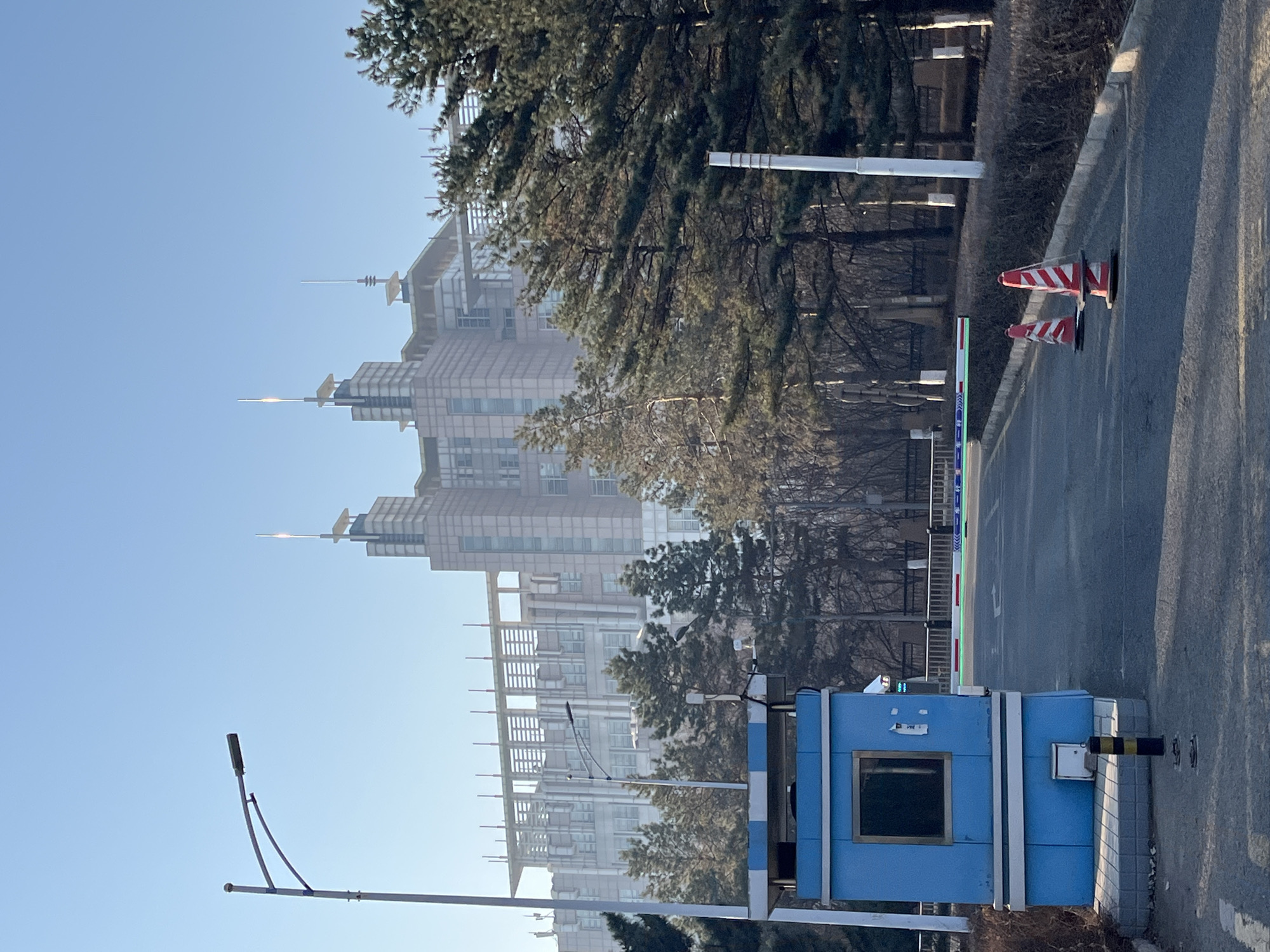 The image depicts an entrance to a property or facility in Changchun City, China. A small, light blue guard booth sits adjacent to an asphalt road. A gate arm, likely for controlling vehicular access, is positioned above the booth. Two red and white striped traffic cones are visible on the road. In the background, several high-rise buildings with distinctive spire-like architectural features are visible, partially obscured by trees. A streetlight is positioned in the foreground, left of the booth. The sky is clear and blue. No people are visible in the frame.