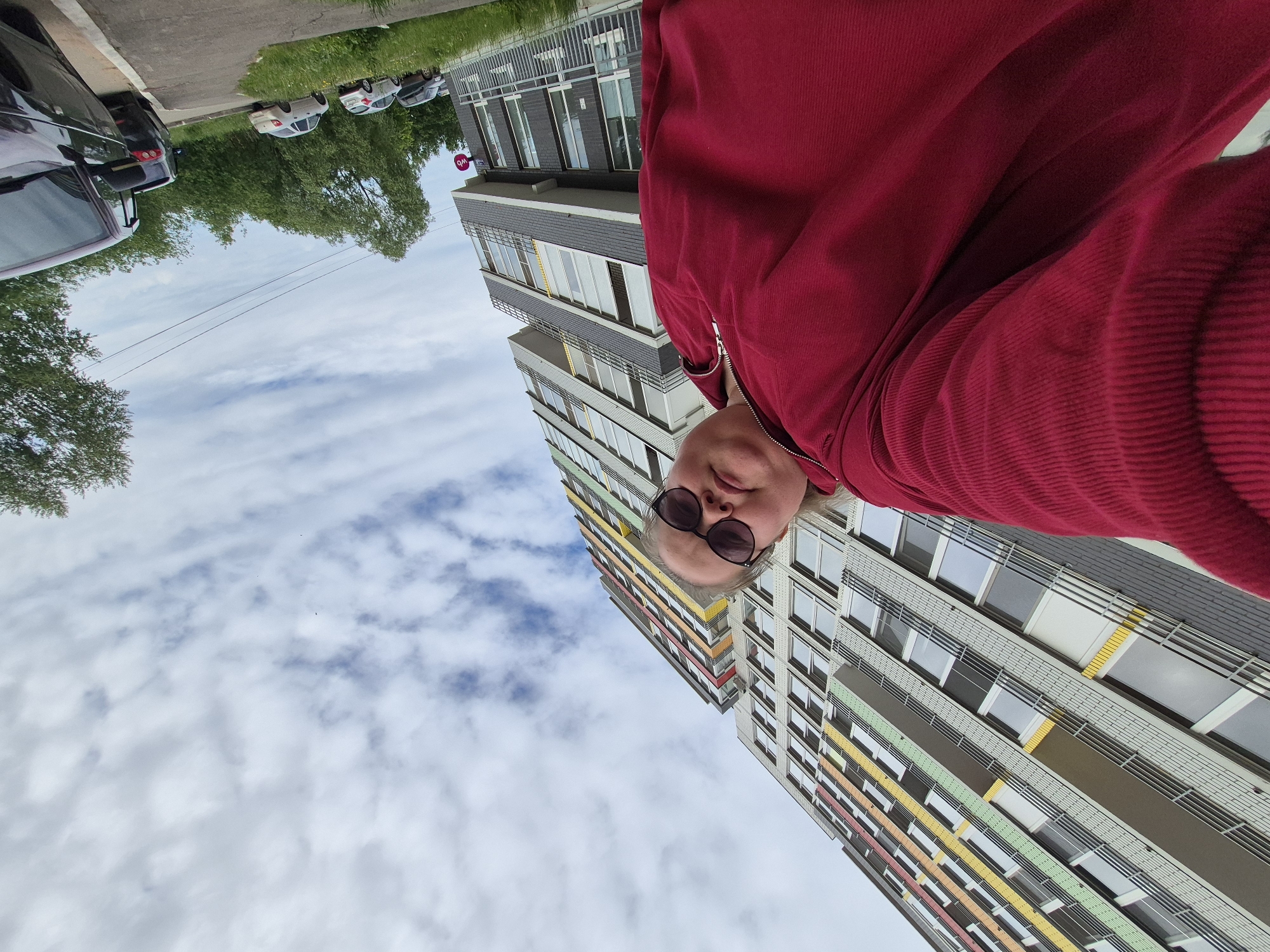 The image captures an outdoor, low-angle selfie featuring a person in the foreground and a multi-story residential building in the background. The person, positioned to the left and center, wears a red corduroy-textured long-sleeved top with a zippered collar and dark-tinted sunglasses, looking directly at the camera.
The prominent structure in the background is a tall residential building with numerous windows and balconies. Its facade is light gray, adorned with horizontal bands of yellow, light green, orange, and red at different floor levels. The building's lower section features darker gray brickwork. A red circular sign bearing the white "wb" logo (Wildberries) is affixed to the ground-floor facade.
To the right of the building, a paved road or parking area is visible, with a strip of green grass separating it from the building. Several vehicles are parked along the road, including a dark-colored SUV in the immediate right foreground. Green trees are present in the middle ground and background on the right. The sky is overcast with scattered white and light gray clouds. The scene is set in an urban area within the city of Chaikovsky, Russia.