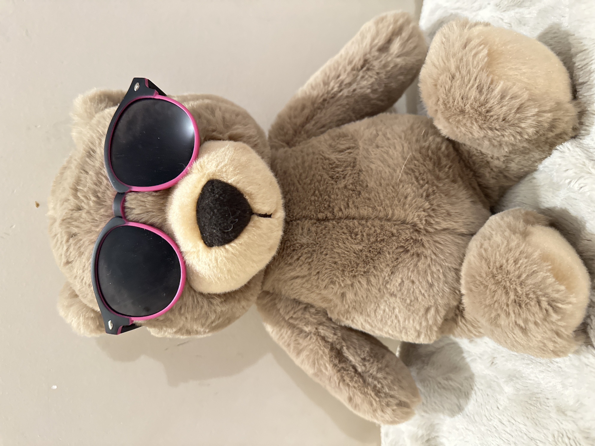 A stuffed teddy bear wearing black sunglasses with pink trim is sitting on a fluffy white blanket. The bear is light brown with a darker brown nose and a light tan muzzle. The background is a plain, lightly textured off-white wall. The image is a close-up shot, focusing on the bear. There is no indication of the location being Tallinn, Estonia.