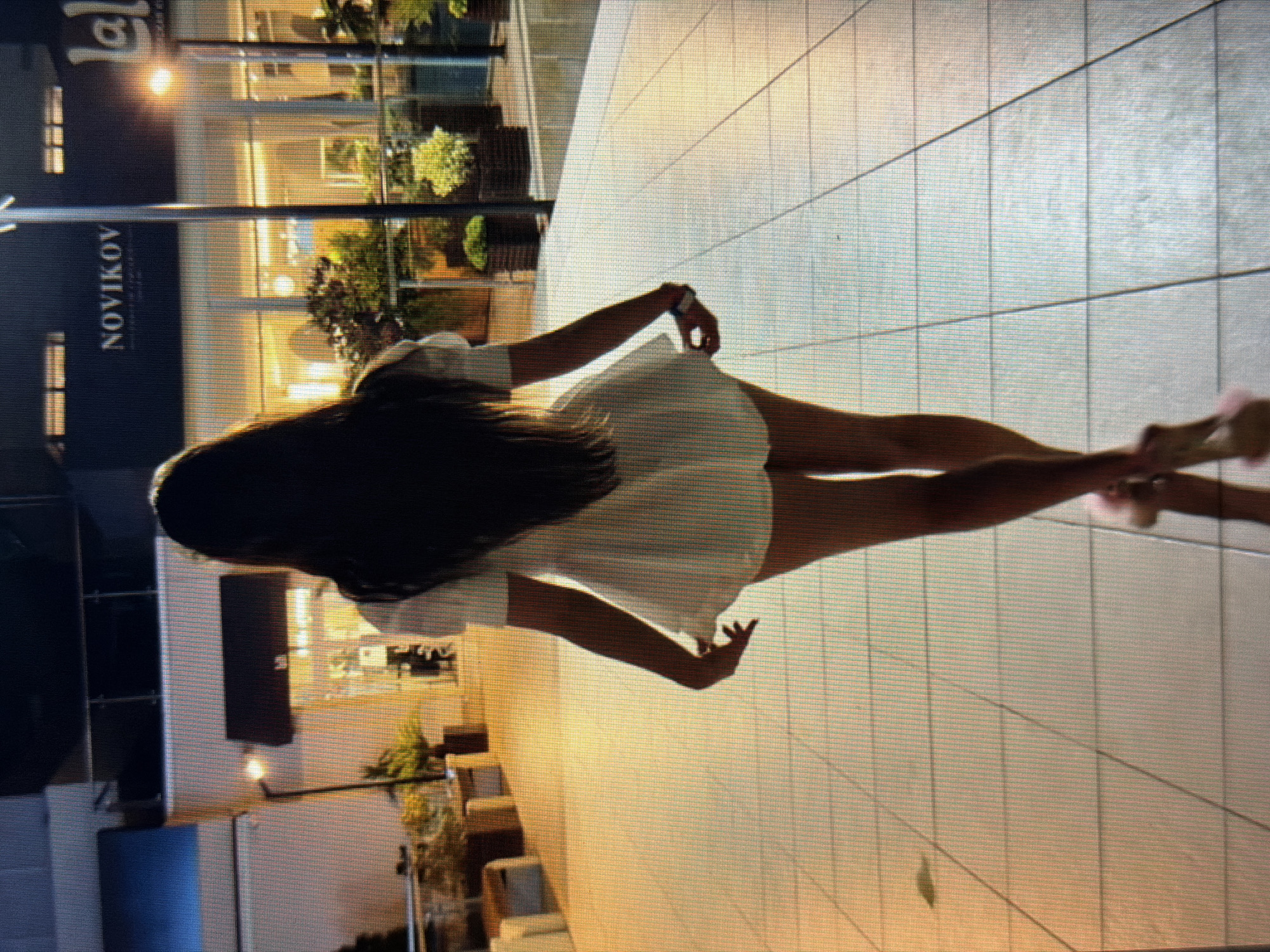 A woman with long dark hair, seen from behind, walks away from the camera on a tiled walkway. She is wearing a short-sleeved white dress that flares out at the hem, and high-heeled shoes. Behind her, a building has the word "NOVIKOV" illuminated in large letters on its dark facade. The building's interior appears brightly lit. The environment suggests an urban setting, possibly a commercial or entertainment district, with some potted plants visible. The lighting is dim, indicating it is likely evening or night. The image does not contain any specific indicators placing it in Sirius, Russia.