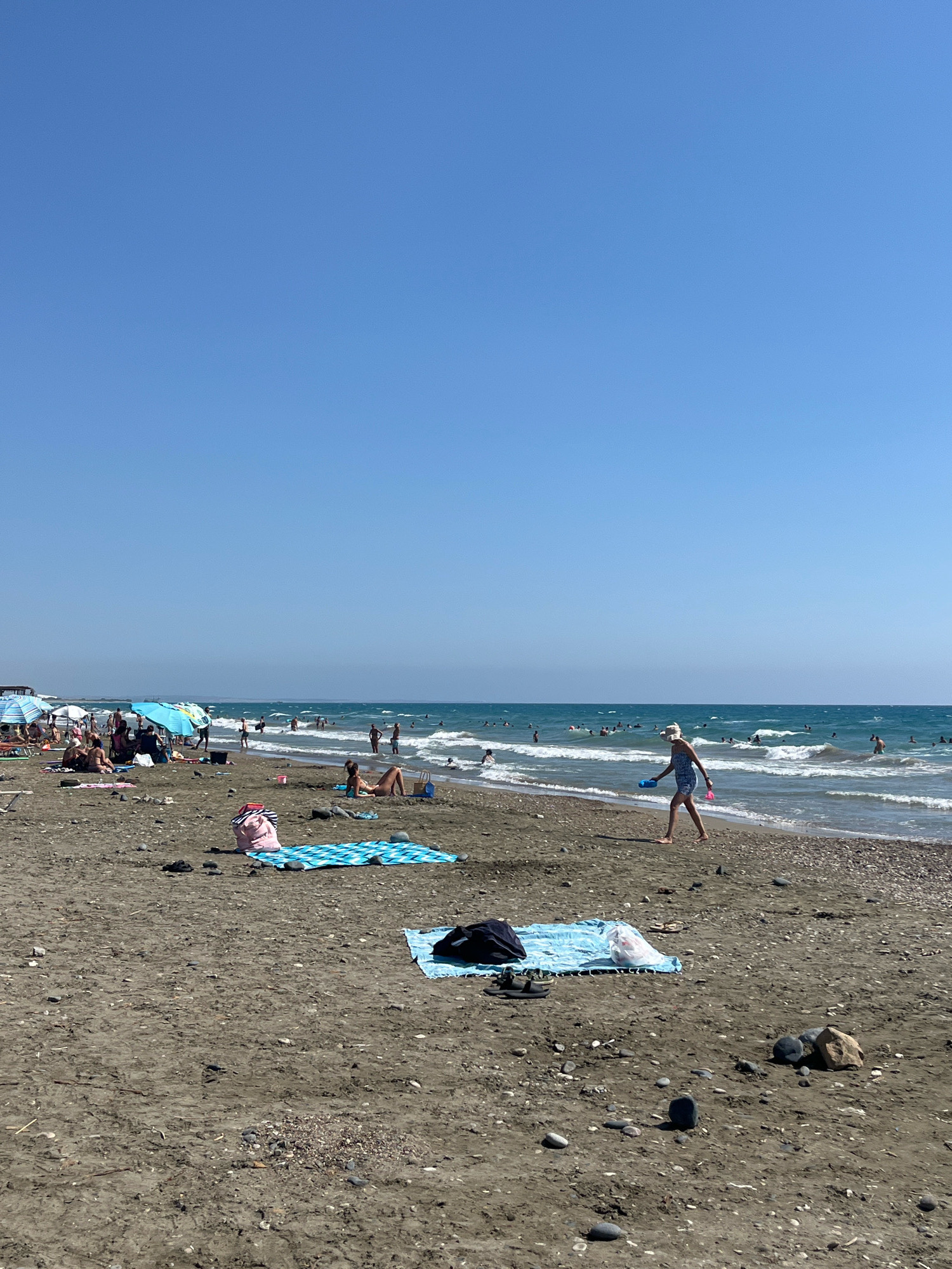 The image depicts a crowded beach on a sunny day, with clear blue skies overhead. The beach is sandy, with some pebbles scattered across the surface. In the foreground, a blue and white striped towel is laid out on the sand, with a pink beach bag on top. Further back, other beach towels and bags are visible.
Numerous people are enjoying the beach. Some are sunbathing on towels, while others are wading or swimming in the turquoise sea. Beach umbrellas, some blue and white, provide shade for groups of beachgoers. The water appears to be relatively calm with small waves breaking near the shore.
In the middle ground, a woman wearing a white hat and a patterned dress is walking along the water's edge, holding blue and pink toys. Other individuals are visible in the water and on the beach, scattered across the scene. The overall impression is of a lively and popular beach destination. The location is identified as Episkopi beach, Limassol, Cyprus.