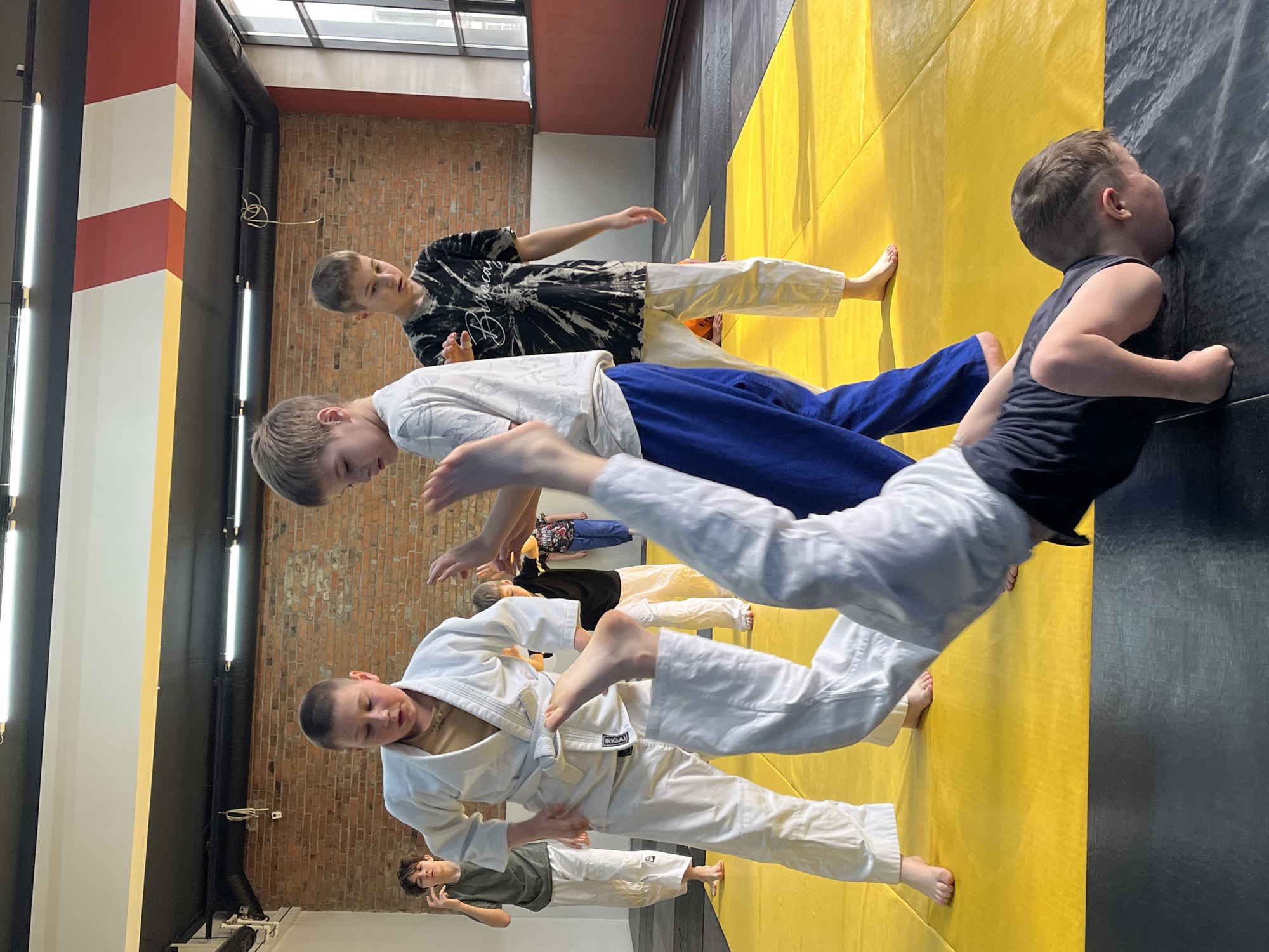 The image depicts a group of young people in what appears to be a martial arts training session in an indoor setting, possibly a gym or dojo. The walls are painted with black, red, and yellow stripes, and there is exposed brickwork visible. A line of young people wearing martial arts uniforms (gi) are practicing a falling or rolling technique on a yellow mat. One person stands by the wall. The location is identified as Krasnoyarsk, Russia.