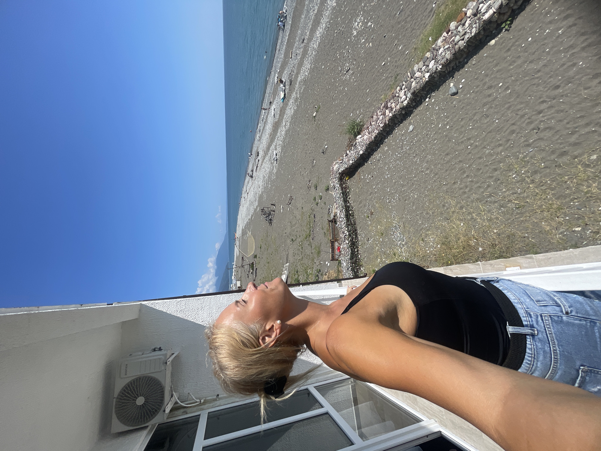 A woman with blonde hair tied back in a ponytail and wearing a black tank top and blue jeans stands on a balcony. She is looking up at the sky with her eyes closed and her face tilted towards the sun. The balcony overlooks a beach with dark sand and pebbles. The sea is visible in the background, with a clear blue sky above. In the distance, mountains can be seen. An air conditioning unit is mounted on the wall of the building to the left of the woman.