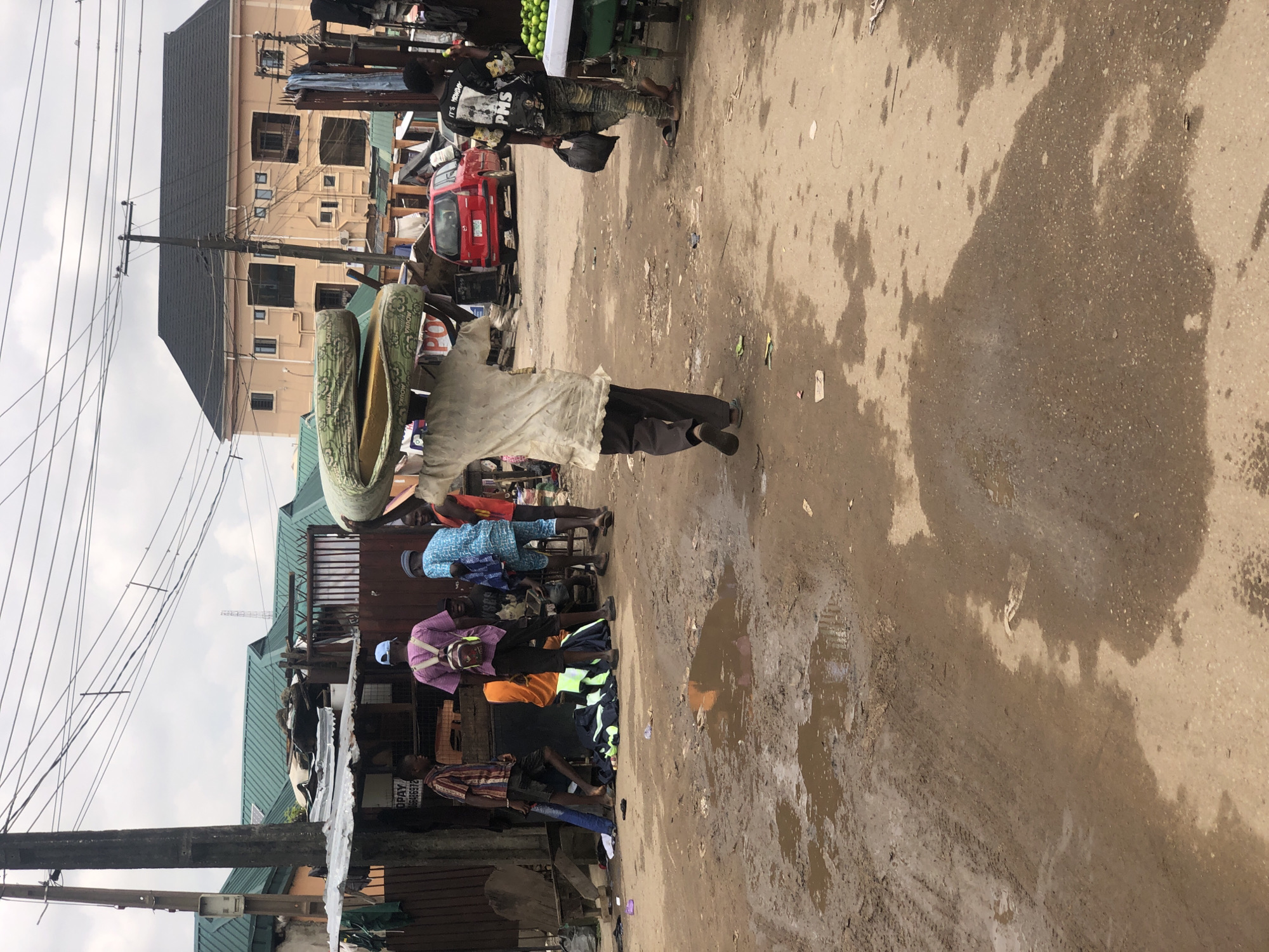 A man, viewed from behind, walks across a muddy, uneven road in Lagos, Nigeria, carrying a large, folded mattress on his head. The mattress is patterned with green and yellow and has writing on it that is difficult to read. The man is wearing a loose, light-colored shirt and dark trousers. The street is lined with stalls and informal shops, some with corrugated metal roofs. In the background, there is a large, light-colored building with a black roof, typical of residential structures in Lagos. Other people are visible in the scene, including a man wearing a purple shirt and backpack, and a child in a blue and white patterned garment. A red SUV is parked on the side of the road. Puddles of water and debris are scattered across the ground, indicating recent rain or poor drainage. The overall impression is of a bustling, commercial street in a densely populated urban environment.