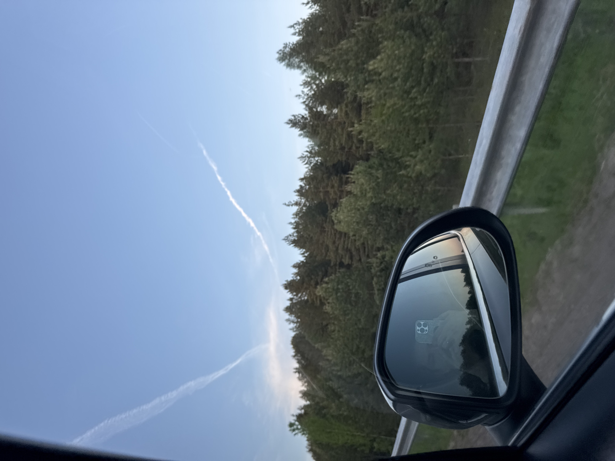 A daytime shot taken from the interior of a moving vehicle captures a view through the car window. A contrail is visible in the upper left sky, with a line of dense trees and a guardrail on the right. A side-view mirror reflects the surrounding scenery. It is not possible to identify the specific location within the city of Bormist, Russia.