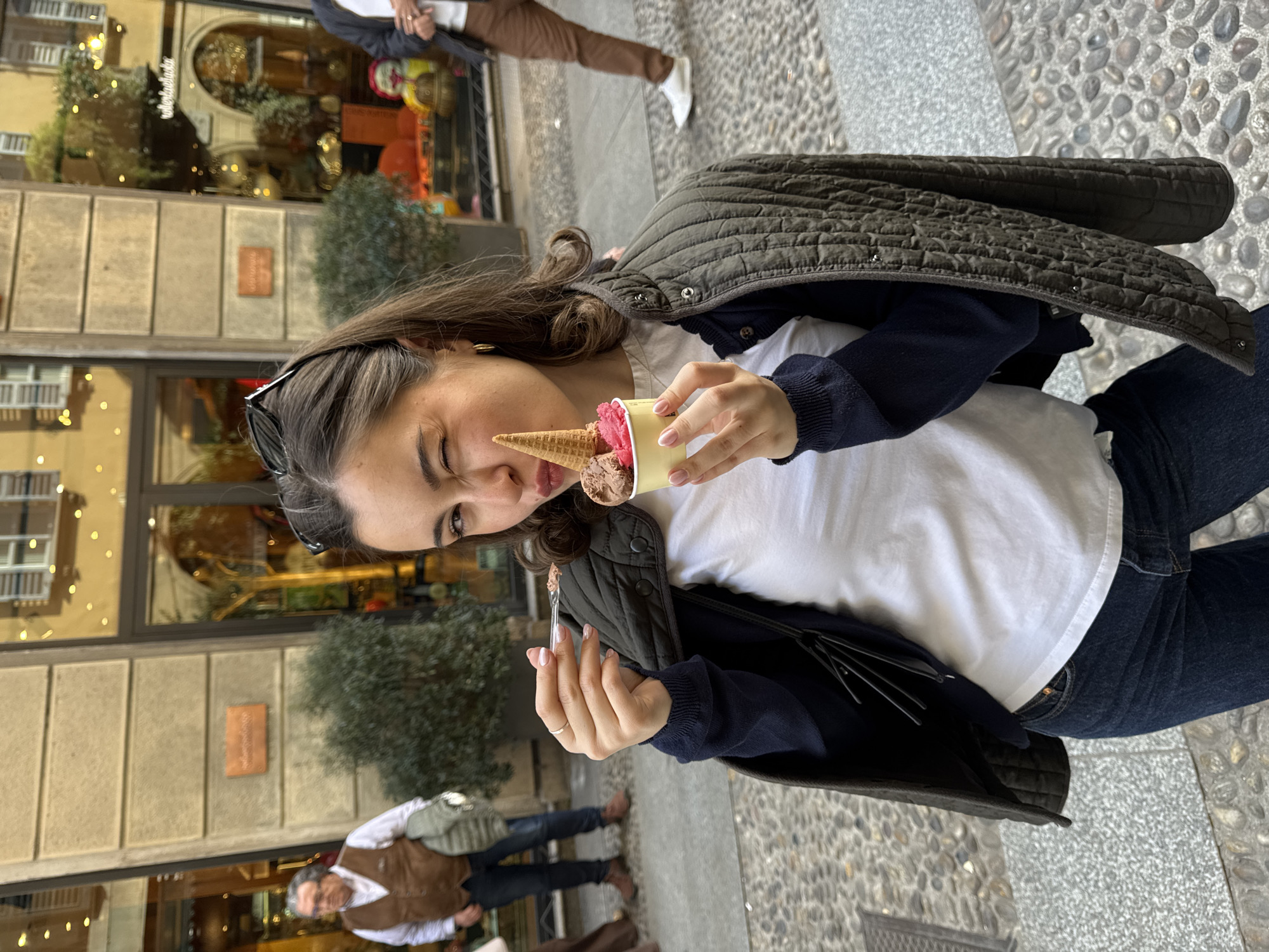 A young adult female is centered in a daytime street scene in Milan, Italy, specifically in front of Roberta Balaska Cioccolato Artigianale, identifiable by signage on the building facade. She wears a white t-shirt, a dark blue long-sleeved top, dark blue jeans, and an open dark olive green quilted jacket draped over her shoulders. Sunglasses are perched on her head, and she has gold hoop earrings and a ring on her left ring finger. The woman is winking with her right eye and pursing her lips while holding a yellow paper cup containing two scoops of gelato (one dark brown, one pink) topped with a small waffle cone in her right hand. A clear plastic spoon, also held by her left hand, is positioned near her mouth. The background features a light-colored stone building with large display windows, reflecting interior lights and showcasing shop items, including foliage in planters outside. The ground is a cobblestone pavement. To the far left, an older male in a brown vest and white shirt walks away. To the far right, a male in brown trousers and white sneakers also walks away, partially visible.