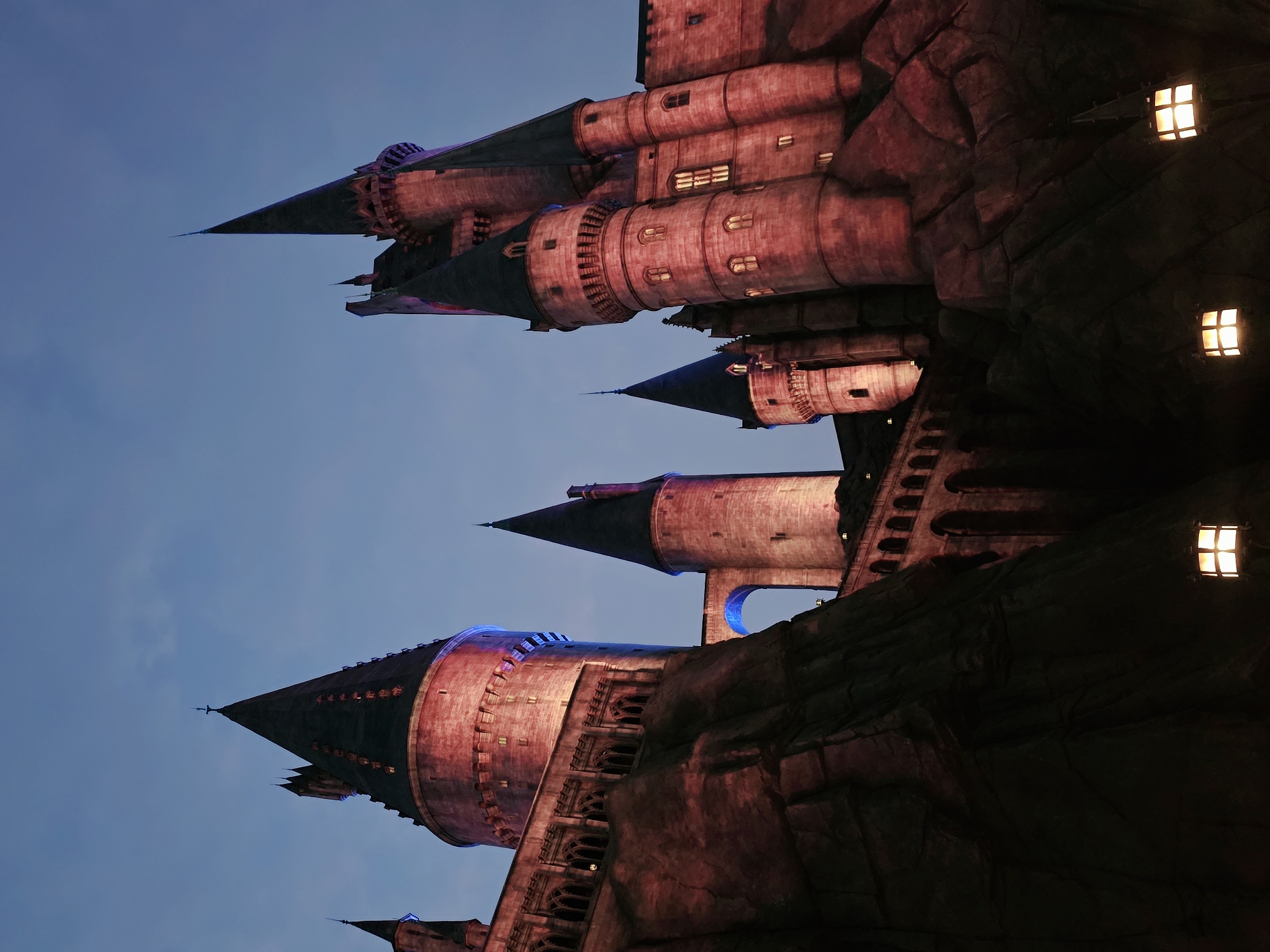 This media file features a nighttime, exterior shot of a medieval-style castle structure. The castle is illuminated with warm red and purple lights, casting dramatic shadows and highlighting its architectural details. Multiple conical towers with dark roofs rise against a clear, dark blue twilight sky. Small, glowing windows are visible on the towers, suggesting occupancy. The castle appears to be built into a rocky, cliff-like formation, adding to its imposing presence. The scene evokes a sense of fantasy and grandeur, possibly related to a theme park or a themed attraction. The lighting and clear sky indicate it is likely evening or early morning. There is no visible text in the image.