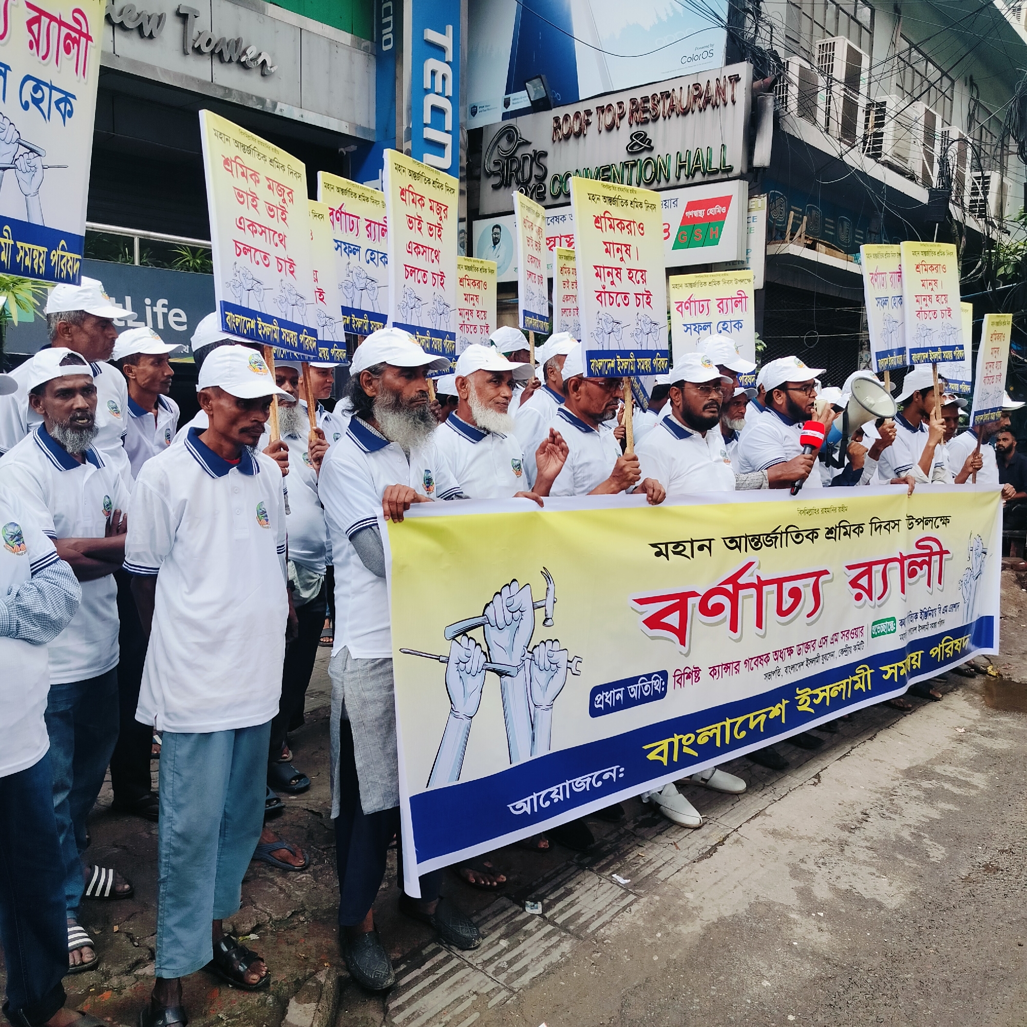 The image depicts a daytime rally or protest, likely in an urban setting, featuring a group of men, mostly middle-aged to elderly. They are uniformly dressed in white polo shirts with a logo on the left chest and white baseball caps. The participants hold numerous placards and a large banner, indicating an organized event.
The placards display slogans in Bengali, such as "May the colorful/grand rally be successful," "Workers and laborers, brothers, want to walk together," and "Workers are also human. We want to live as humans." Many placards also feature an illustration of several hands holding various tools, including a hammer, screwdriver, and wrench, symbolizing manual labor.
The large yellow banner, prominently held at the front, also features this tool illustration. The text on the banner begins with "বিসমিল্লাহির রাহমানির রাহীম" (In the name of Allah, the Most Gracious, the Most Merciful) and states "মহান আন্তর্জাতিক শ্রমিক দিবস উপলক্ষে বর্ণাঢ্য র্যালী" (Colorful/Grand Rally on the occasion of Great International Workers' Day). It identifies the organizer as "বাংলাদেশ ইসলামী সমন্বয় পরিষদ" (Bangladesh Islamic Coordination Council) and mentions a chief guest, "বিশিষ্ট ক্যান্সার গবেষক অধ্যক্ষ ডাক্তার এস এম সরওয়ার" (Eminent Cancer Researcher Principal Dr. S M Sarwar).
In the background, multi-story buildings are visible, adorned with various signs including "New Tower," "TECHNO," and "ROOF TOP RESTAURANT & CONVENTION HALL." Numerous electrical wires are visible cascading down the buildings. The street appears damp, suggesting high humidity or recent rain. The expressions of the participants are generally serious and focused, conveying the gravity of their cause.