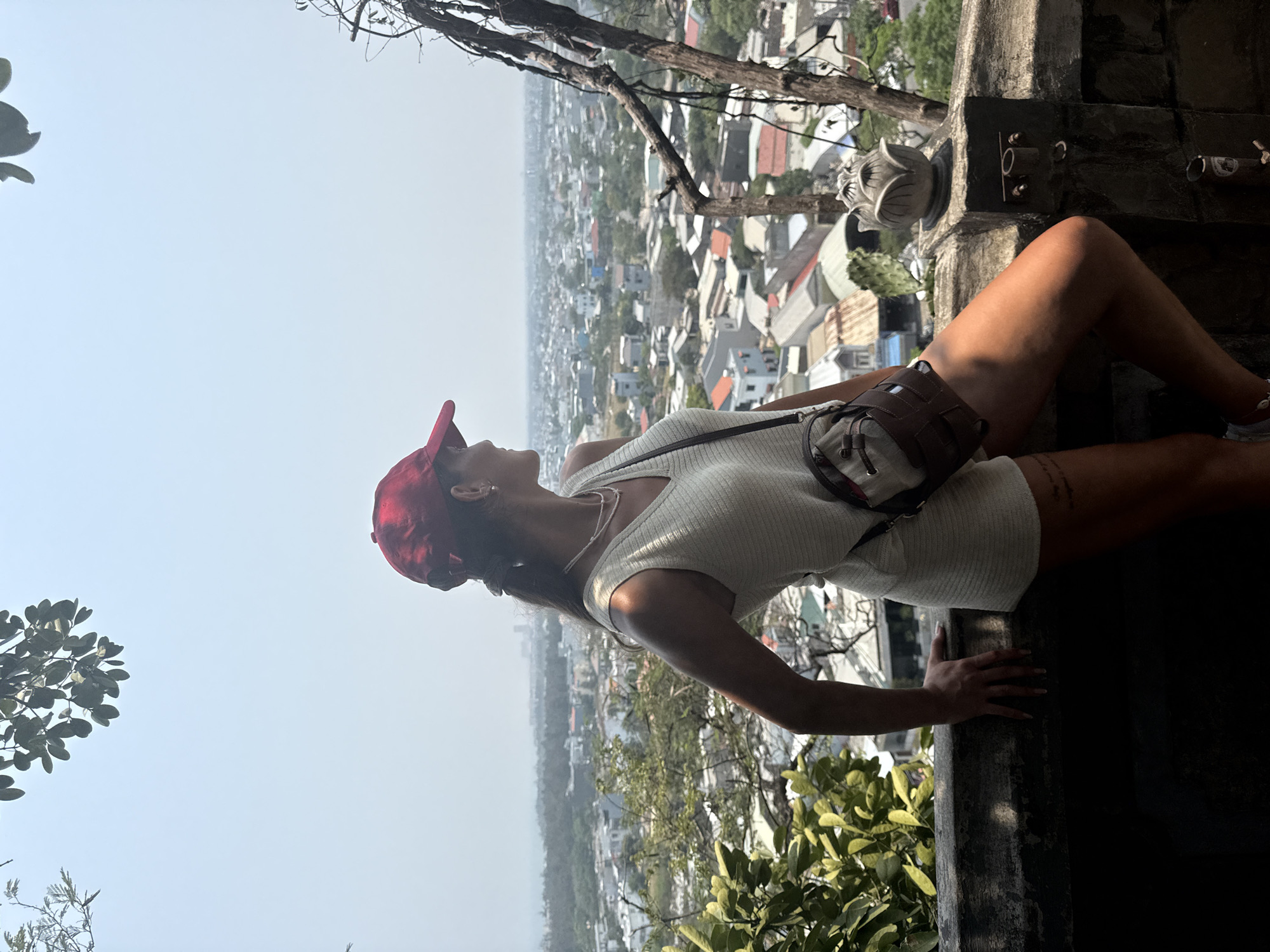 A woman wearing a red cap, a white ribbed dress, and a cross-body bag is leaning against a concrete ledge, overlooking a densely populated urban area in Đà Nẵng, Vietnam. The city sprawls behind her, with buildings and houses visible in the hazy background. A tree branch is visible above her. A lotus flower statue sits to the right of her, atop the ledge. Vegetation grows below the ledge.