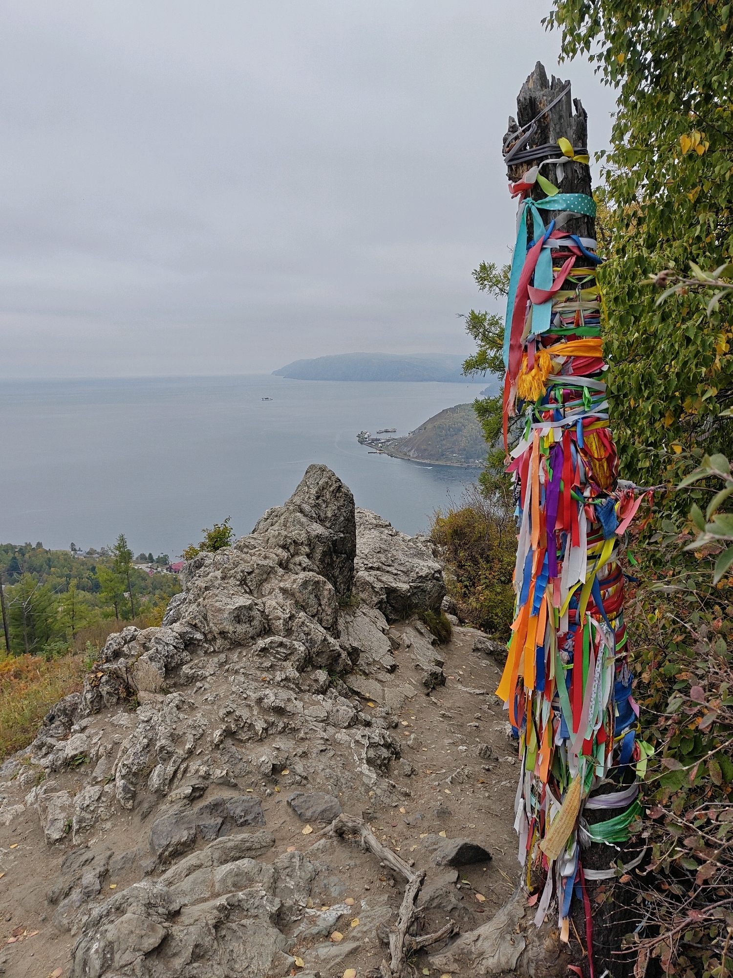 This image captures a natural landscape scene with a cultural element at a location near Nikola, Russia. In the foreground, a weathered, dead tree stump stands vertically, heavily adorned with numerous colorful ribbons of various lengths and hues. The ribbons are tied around the stump in a festive, ritualistic manner. Adjacent to the stump, a rocky, uneven path winds through the foreground, with scattered stones and fallen leaves.
Behind the stump and path, the terrain slopes downward, revealing a rocky outcrop in the mid-ground. Further in the distance, a vast body of water, likely a lake, stretches towards the horizon. The water appears calm under an overcast sky. Along the far shore, a hilly landscape is visible, with a small settlement nestled at the base of a peninsula jutting into the water. A body of water, possibly a bay or inlet, is visible to the right of the peninsula, with a ship or boat on its surface. The overall atmosphere is subdued, indicative of an autumn day. The image does not depict any people or animals.