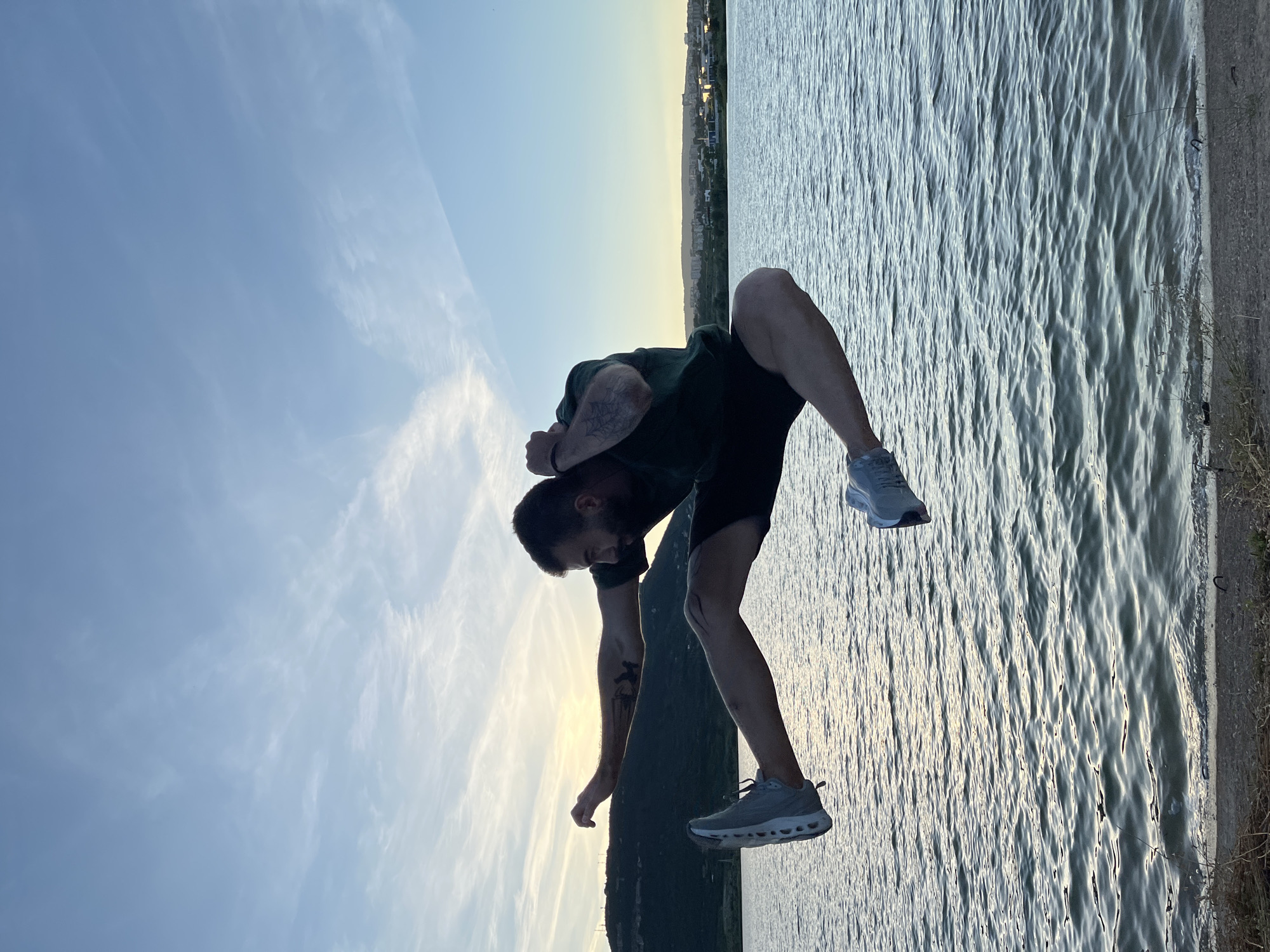 A man with a beard and tattoos on his arms is captured mid-air in a dynamic jump near a body of water. He is wearing a dark green t-shirt and black shorts, with gray athletic shoes. His left arm is extended forward, while his right arm is bent upwards towards his head. His left leg is extended forward and parallel to the ground, with his right leg bent at the knee. The background features a serene lake with gentle waves and a distant shoreline with rolling hills and a city skyline under a pale blue sky with wispy clouds. The lighting suggests it is either sunrise or sunset. The image appears to have been taken from a slightly low angle, emphasizing the man's airborne pose. This scene could be located at the Shumen Plateau, which overlooks the city of Shumen, Bulgaria, a region known for its natural beauty and recreational opportunities.