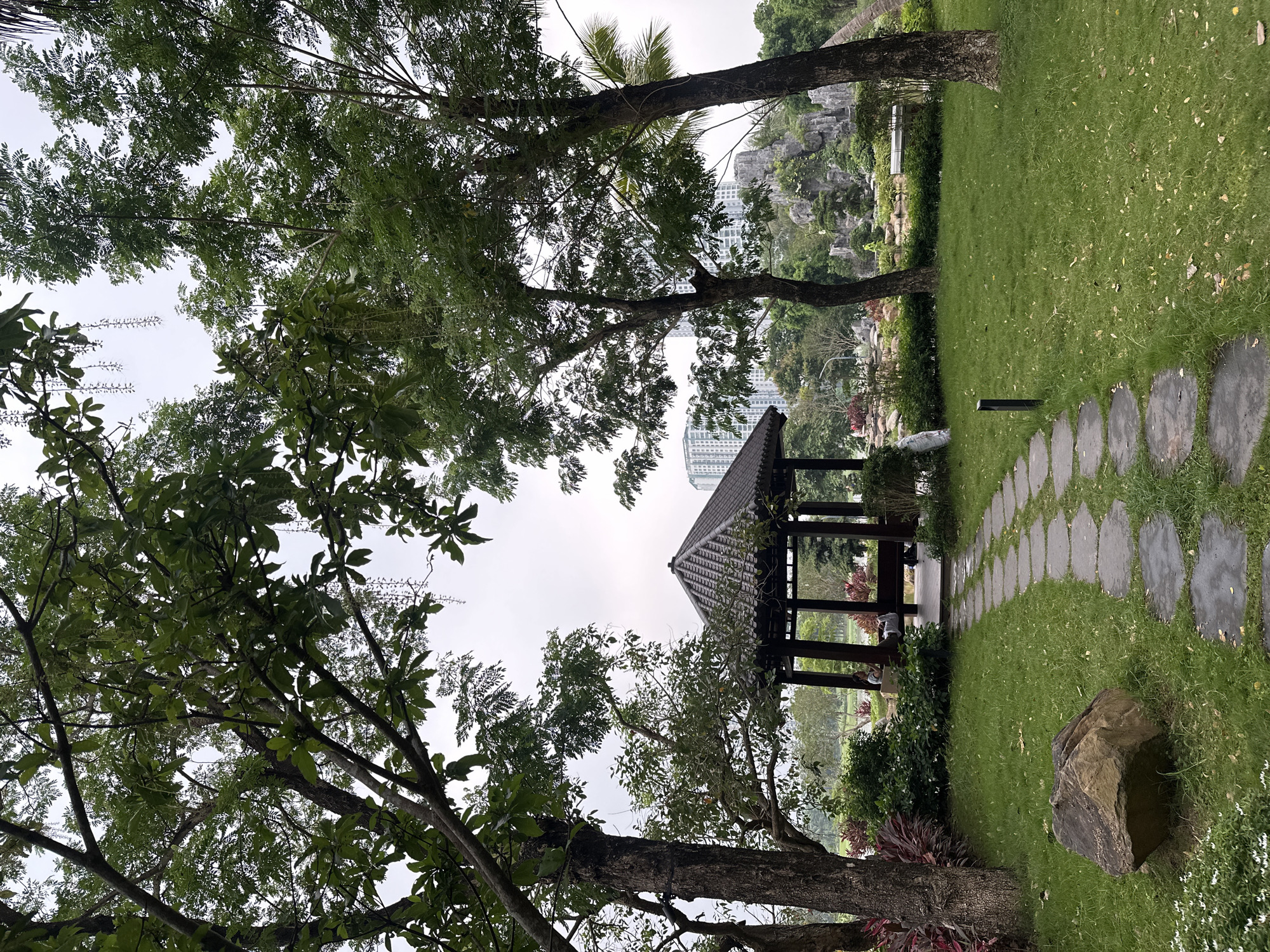 In the city of Phụng Công Commune, Vietnam, a stone pathway winds through a lush green lawn, leading to a dark wooden gazebo with a grey tiled roof. The gazebo is open on the sides, revealing two people seated inside. Tall trees with dense green foliage frame the scene, with branches extending towards the overcast sky. In the background, rocky formations rise amidst more trees and vegetation, with modern buildings visible in the distance. A large, dark boulder sits on the grass to the left of the path.