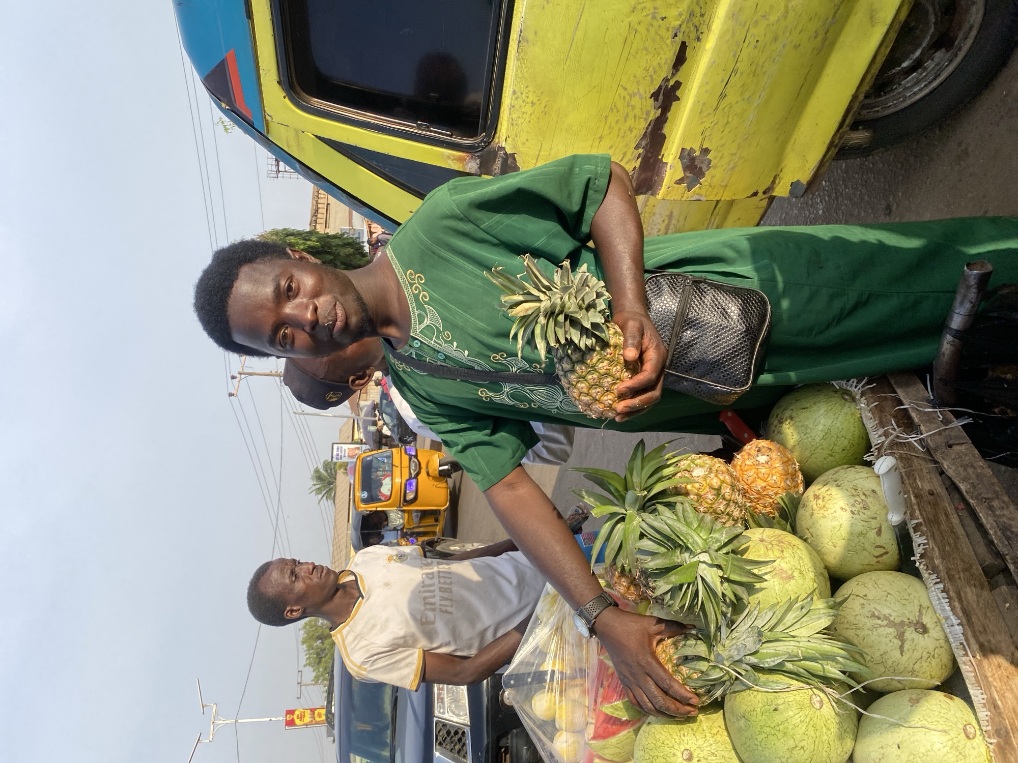 The scene depicts an outdoor street market in Sabon Tasha, Nigeria, under a clear, bright sky. In the foreground, a male with dark skin and a short, dark afro hairstyle, wearing a green long-sleeved traditional tunic with embroidered patterns and a black textured crossbody bag, is positioned centrally. He has a nose piercing on his left nostril, is looking towards the viewer, and holds two small pineapples in his left hand. His right hand is extended towards a wooden fruit cart in front of him, which is laden with watermelons and pineapples. To the right of the central figure, a yellow vehicle panel with extensive paint peeling and rust is visible.
Behind the fruit cart, a second male with dark skin, wearing a white t-shirt with "Emirates FLY BETTER" and a Real Madrid logo, stands partially obscured, looking to his left. A dark SUV is partially visible behind him. Further into the midground, a yellow motorized rickshaw (keke Napep) is present on a paved street. The background features utility poles, overhead power lines, and indistinct buildings, indicating an urban setting. A third individual's head, wearing a dark cap, is partially visible behind the central figure.