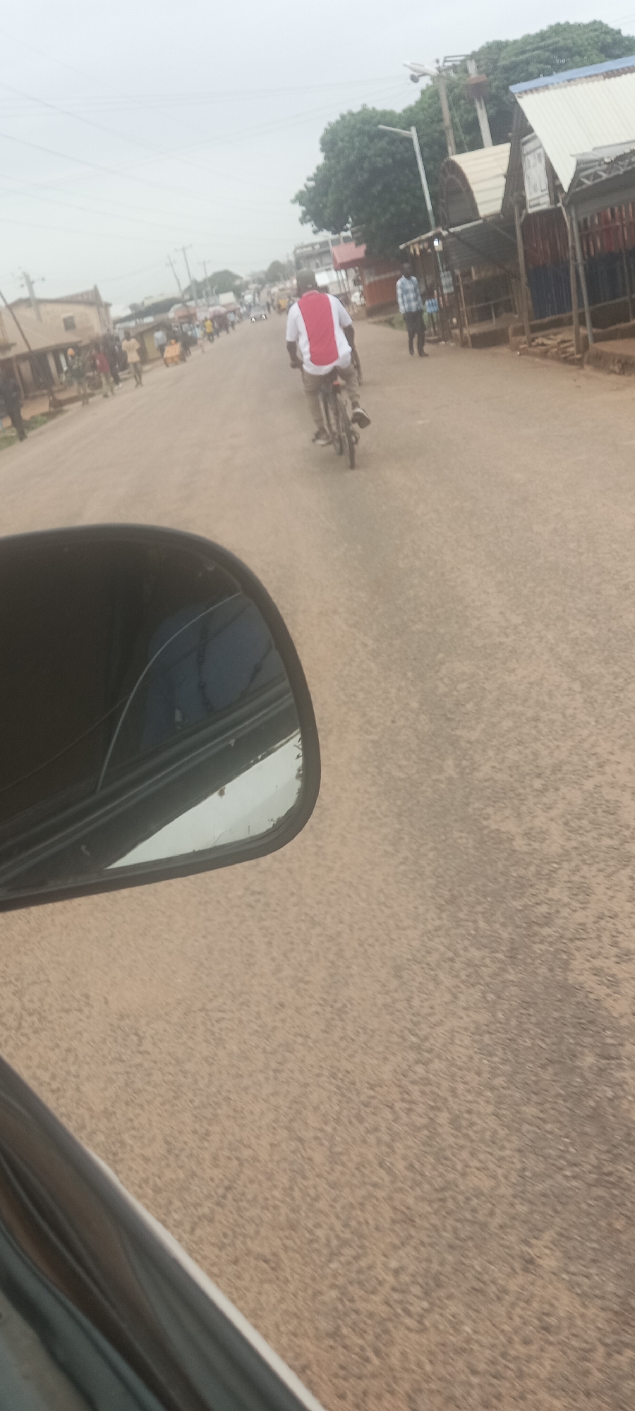 The scene is a street in Barakin, Nigeria. A man wearing a white shirt with a red stripe is riding a bicycle down a dusty road. Several people walk along the sides of the street near buildings. The photo is taken from inside a car, with the side mirror visible in the foreground.