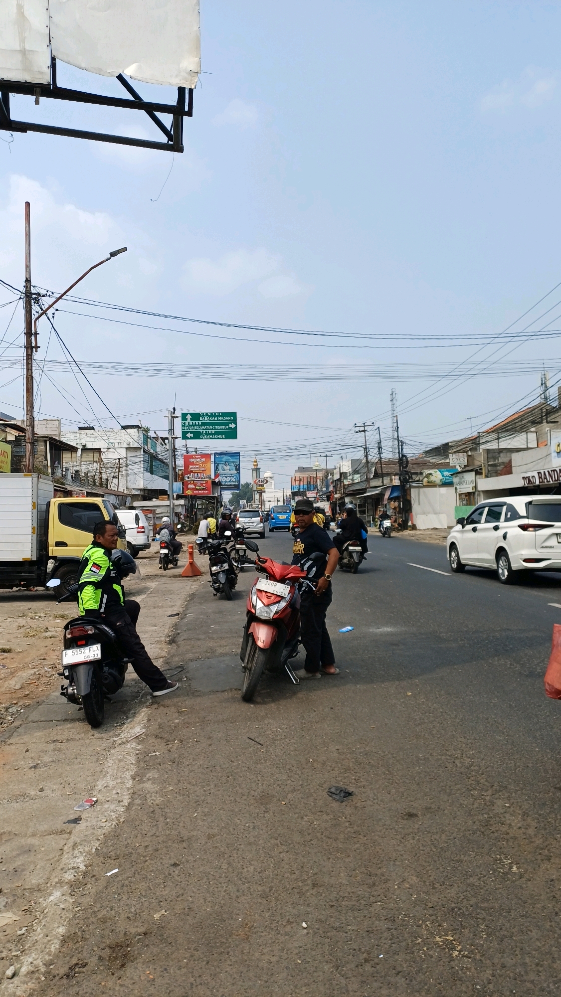 The image captures a street scene in Citeureup, Indonesia. In the foreground, two men are standing near motorcycles on a paved road. One man, wearing a bright green and yellow jacket, is seated on a black motorcycle. The other man, dressed in a black t-shirt and pants, stands next to a red motorcycle. Several other motorcycles and a white car are visible on the road, suggesting traffic. The background features buildings, shops, power lines, and a green street sign indicating directions to various locations, including "SEKTOR BARAK MADANG" and "CIBINONG". The sky is partly cloudy. The overall scene depicts a typical urban street environment with ongoing traffic and pedestrian activity.