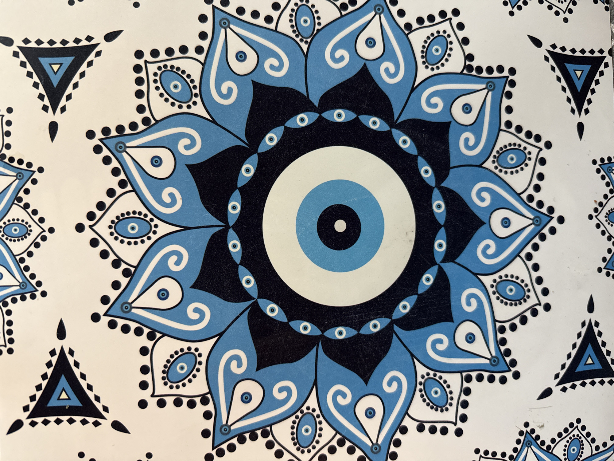 The image features a pattern dominated by a circular, stylized evil eye design in shades of blue, black, and white. The central eye is surrounded by concentric rings of color and decorative elements. Radiating outwards are stylized petals with white scroll-like decorations against a blue background, some containing smaller eye-like motifs. The petals are edged with black dots. The background consists of white triangles outlined in black, with smaller triangular motifs inside, each with a small blue eye in the center.
There is no information to suggest any location in the city of Pleven, Bulgaria.