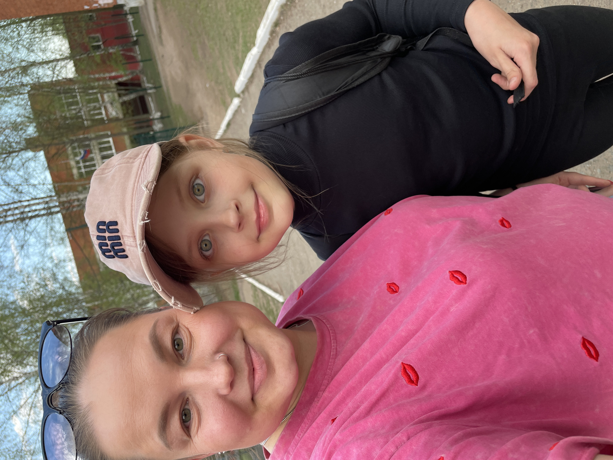 Two females are taking a selfie outdoors. The individual in the foreground wears a pink t-shirt with embroidered lips and sunglasses resting nearby. The other, presumably a child, is behind and to the side. The child wears a cap. The background includes an unidentifiable building, trees, and a grey surface, suggesting an urban or suburban outdoor setting. The Russian flag can be partially seen on one of the buildings. Based on the location of the flag and buildings, the location is Izhevsk, Russia.