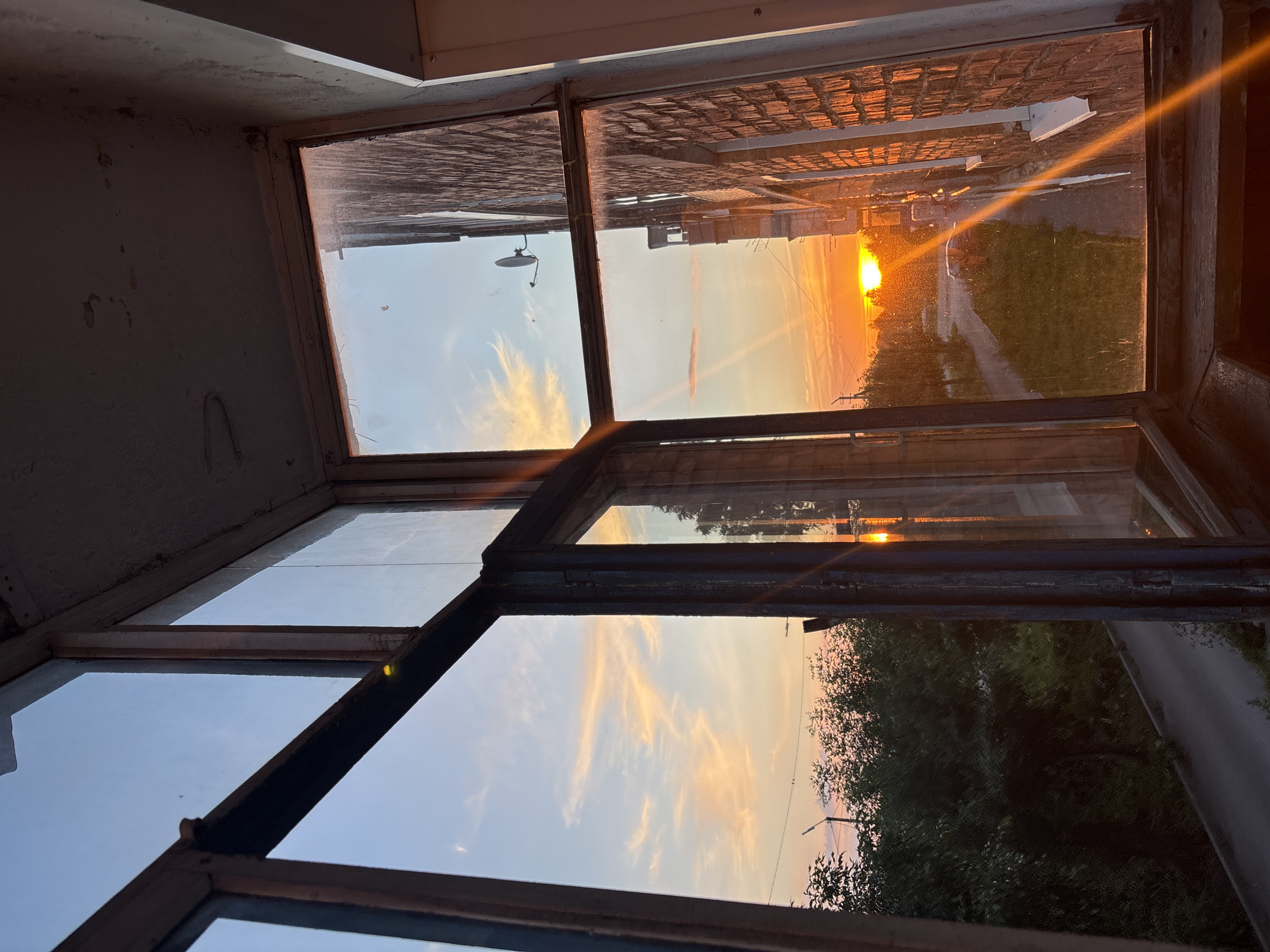 This is a photograph taken through a multi-paned window, likely from an interior space looking outwards. The perspective is at an angle, making the scene appear somewhat tilted. The window frames are dark and appear worn, with some dirt and debris visible on the panes and frames.
The scene depicted outside the window is a sunset. The sun, a bright orange disc, is low on the horizon, casting a warm, golden light across the sky and landscape. Wispy clouds are visible, tinged with orange and pink from the setting sun. Beyond the clouds, the sky transitions to a soft blue.
In the foreground of the scene, through the different panes of the window, are reflections and partial views of the surrounding environment. On the left side of the frame, the interior wall is visible, with textured paint and what appears to be some water staining. Through the upper right pane, a brick wall of a building is visible, bathed in the sunset's glow. A path or road is also visible, running through a grassy area. To the right, dense green trees are visible, their foliage silhouetted against the brightly lit sky. A road or path is also discernible among the trees.
The overall mood is serene and peaceful, evoked by the warm colors of the sunset and the gentle sky. The time of day is clearly evening, as indicated by the sunset. The location cues, such as the brick building and the type of vegetation, are consistent with a residential or semi-rural setting. No text is visible in the image. The image appears to be taken in Inskoy, Russia.
