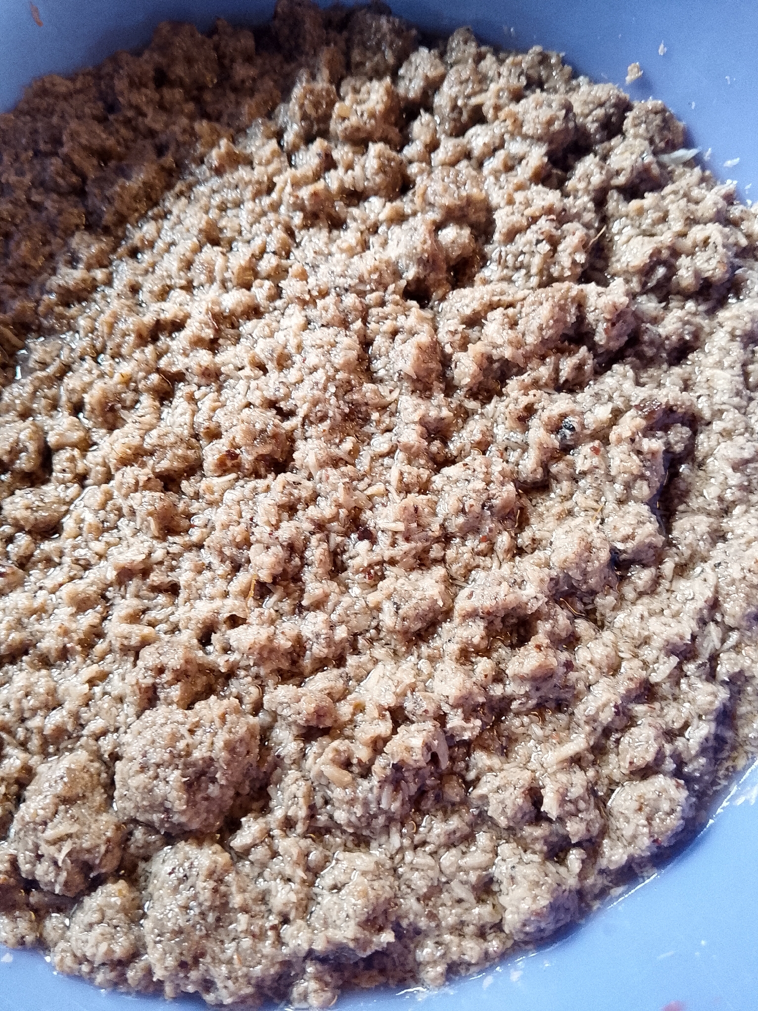 This is a close-up, overhead, outdoor, medium shot of a light blue plastic bowl filled with a moist, ground, meat-like substance. The substance is a light brown to greyish-brown color and has a granular, clumpy texture with some visible liquid. The bowl's edge is visible along the bottom and right sides of the image. The lighting suggests it is daytime, and the overall scene appears to be a food preparation or serving context. There are no identifiable landmarks or elements that can place this image specifically within the city of Lhokseumawe, Indonesia.