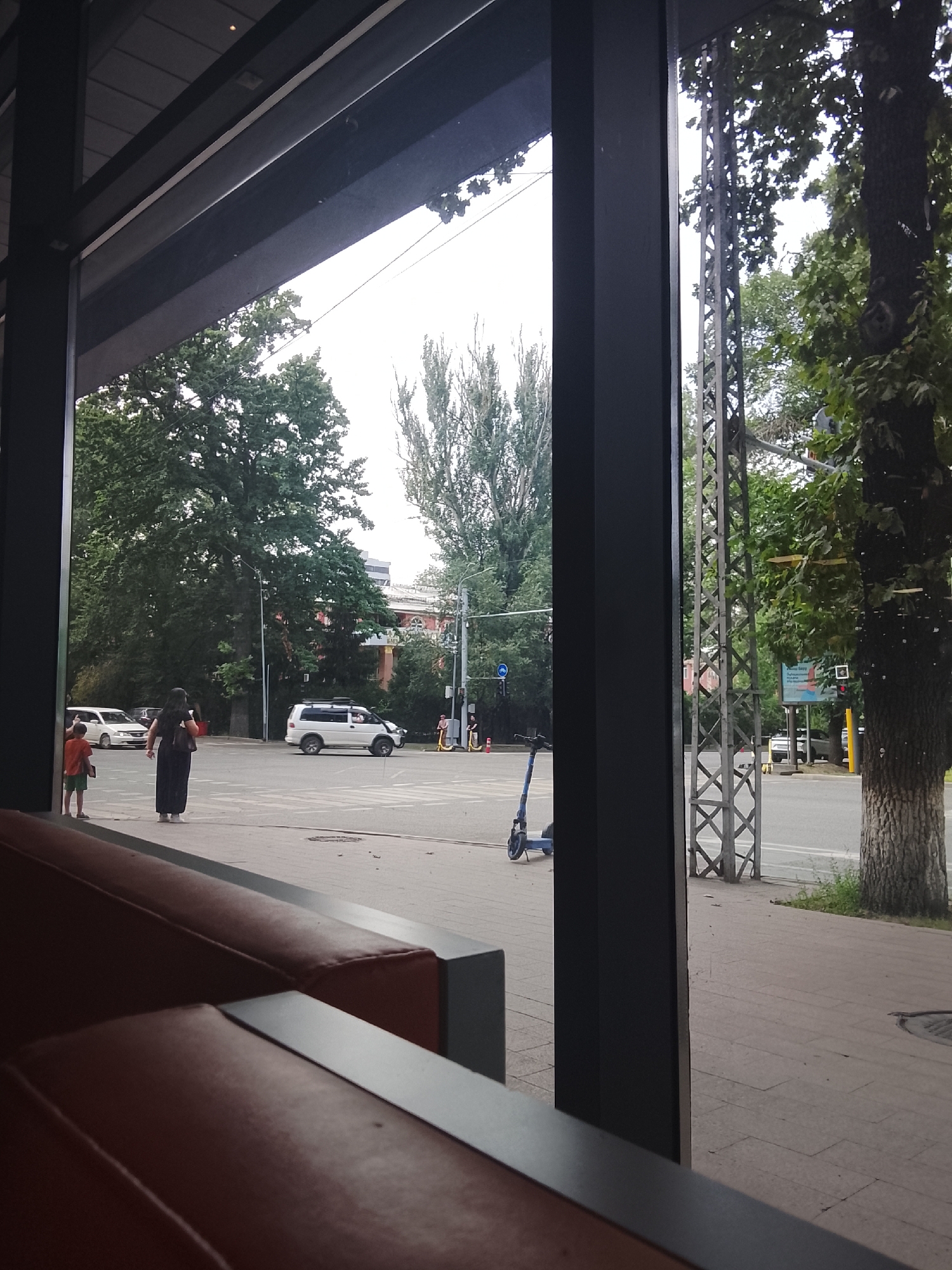 The image depicts an exterior view from what appears to be a cafe or restaurant, looking out onto a street intersection in Almaty, Kazakhstan. In the foreground, out of focus, are reddish-brown upholstered seats. The main scene shows a street with pedestrian crossings and a traffic light.
Several individuals are visible. On the left side of the street, a child in an orange shirt and green shorts stands near a woman in a long, dark dress. Across the intersection, several people are seen near a white SUV and a white van. Two individuals are on yellow electric scooters on the right side of the street.
The environment is urban, with trees lining the street and buildings in the background. A metal tower structure stands prominently on the right side of the image, next to a large tree. The sky is overcast, suggesting a cloudy day. A blue electric scooter is parked on the sidewalk in the lower left portion of the frame. The overall impression is of a typical street scene in a city.