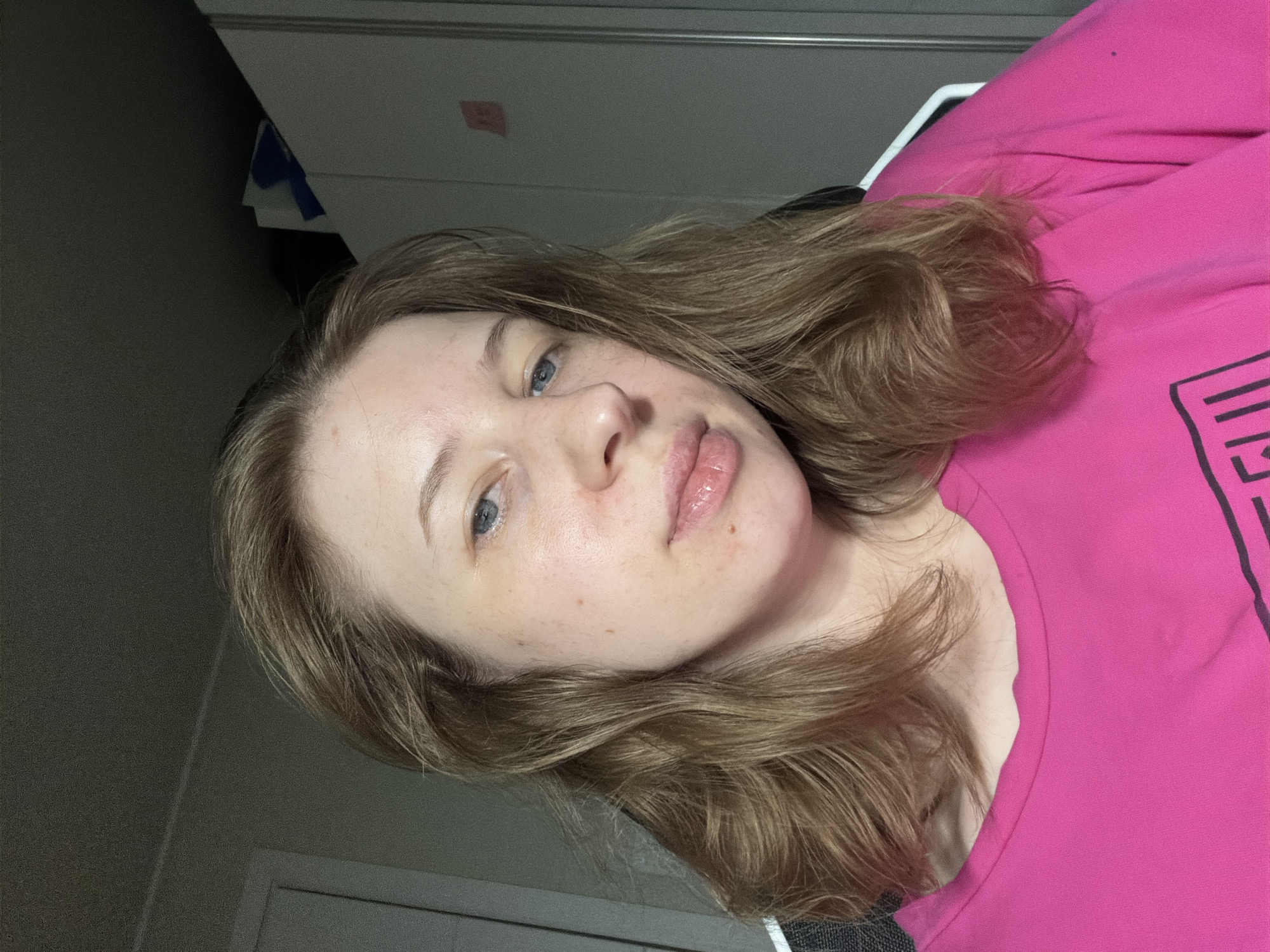 A medium shot photograph shows a young Caucasian woman from the shoulders up, facing the camera. She has light skin with some visible blemishes, light blue eyes, and shoulder-length light brown hair. She is wearing a bright pink t-shirt with a black graphic design visible on the lower right side. The background consists of a plain gray wall and a white cabinet on the right. The lighting appears to be artificial and coming from above and the front. The image was taken indoors in a residential setting. The scene does not provide any specific information about its location within Dongducheon-si, South Korea.