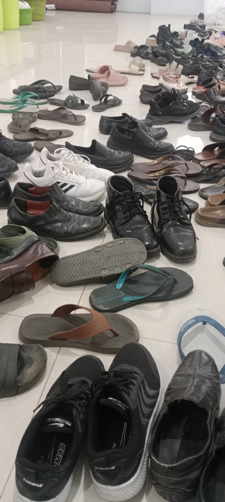 The image depicts a cluttered collection of footwear, primarily shoes and sandals, scattered across a light-colored tiled floor. The scene appears to be an indoor storage area or entryway, possibly in a residence or a shared facility. The items are diverse in style, color, and condition, ranging from athletic shoes and formal leather shoes to various types of sandals and flip-flops. Some items are neatly arranged, while others are piled or strewn about, suggesting a lack of organization. The lighting is ambient, casting soft shadows and highlighting the textures of the different footwear materials. Based on the context provided, this scene is located in the city of Tuban, Indonesia.