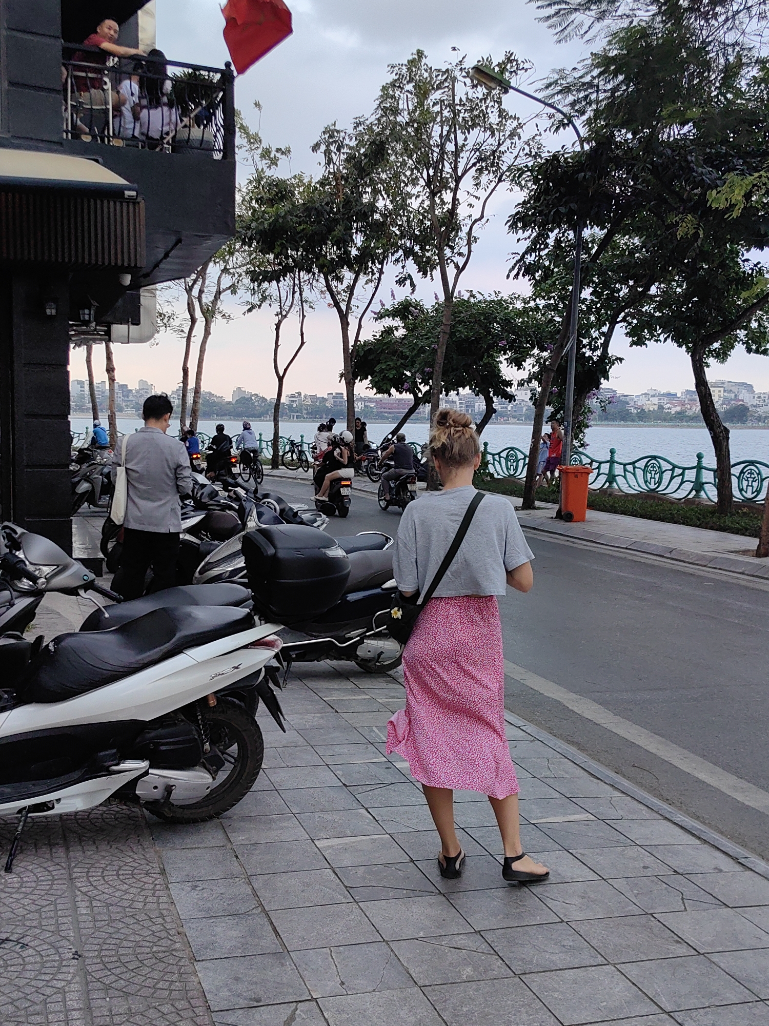 The photograph depicts a street scene in Hà Nội, Vietnam. The focal point is a woman walking away from the viewer on a sidewalk. She has her hair in a bun and is wearing a gray top, a pink floral skirt, and black sandals. A black strap bag is slung across her shoulder.
To the left, there are several parked scooters and a man standing near them wearing a gray suit. Further down the street, other people are seen on bicycles and scooters.
On the right side of the road, a decorative green railing separates the sidewalk from a body of water. Trees line the street.
In the background, buildings are visible across the water. Above the buildings is a cloudy sky.
On the left side of the image, there is a building with a balcony. People can be seen standing on the balcony, and a red flag hangs from it.