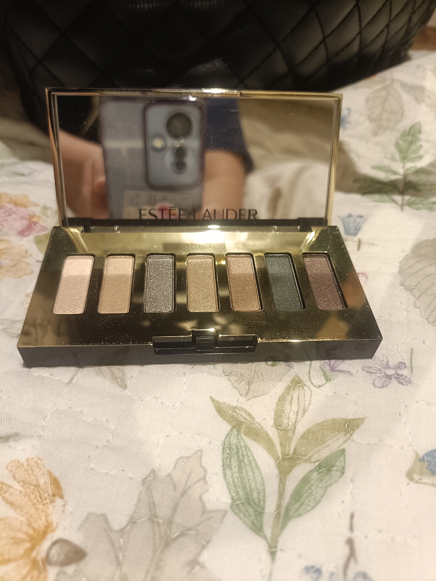 An overhead shot shows an open Estee Lauder eyeshadow palette with eight shades on a quilted bedspread. The palette is gold with a black base and a mirrored lid that reflects a cellphone. The eyeshadow shades, from left to right, are a pale pink, a light beige, a silvery grey, a golden brown, a muted taupe, a dark green, and a deep brown. The bedspread has a floral pattern in shades of green, yellow, and light purple. The setting is indoors, likely a bedroom. There is no indication of the location being in Dubai, United Arab Emirates.