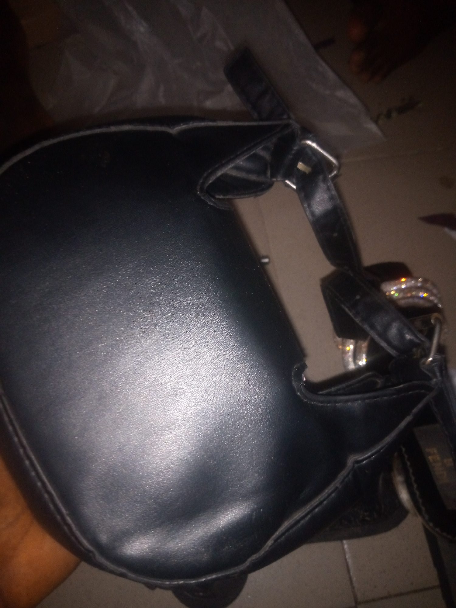 A black handbag is displayed against a dark background. The handbag appears to be made of leather and has a somewhat worn appearance with visible stitching along its edges. A strap is visible attached to the bag. The handbag is resting on a tiled surface. A faint pattern or texture is visible on the surface beneath the bag. In the upper right corner, a person's feet are partially visible, suggesting they are standing or sitting near the handbag. The lighting in the image is dim, creating shadows and highlighting the texture of the handbag. The overall scene appears to be indoors, possibly in a home or retail environment. The context suggests the handbag is a fashion accessory.