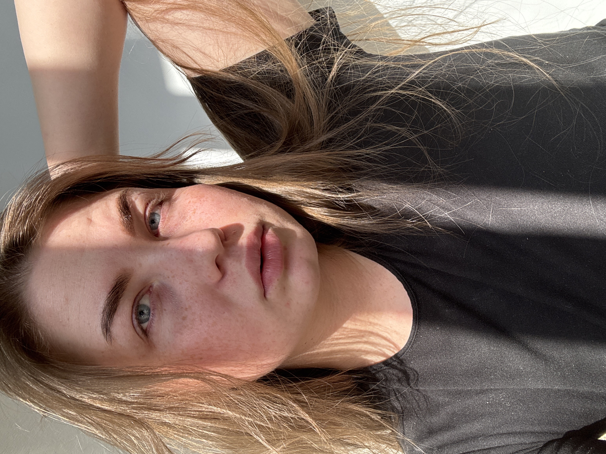 A young woman is depicted in a close-up portrait, oriented horizontally as if lying down or reclining. Her head is positioned to the left, with her body extending to the right. She has light brown or blonde hair, which is spread out around her head and shoulders, with individual strands highlighted by strong sunlight. Her blue eyes are looking directly at the camera, and her lips are slightly parted. Her skin shows visible freckles, particularly across her cheeks and nose.
The woman is wearing a plain black t-shirt or top. Her right arm is visible in the upper left corner of the frame, bent at the elbow.
The scene is illuminated by bright, natural sunlight, likely streaming from a window, which casts distinct shadows across her face, particularly over her forehead and left eye, and on her neck and chest. This creates strong contrasts between illuminated and shadowed areas. The background is a simple, light-colored surface, possibly a wall or floor, contributing to a clean and uncluttered composition.
Her facial expression is neutral and relaxed, suggesting a moment of calm or repose. There is no visible text in the image. The strong sunlight indicates it is daytime, either morning or afternoon. The overall impression is a natural and intimate portrait, enhanced by the play of light and shadow.
